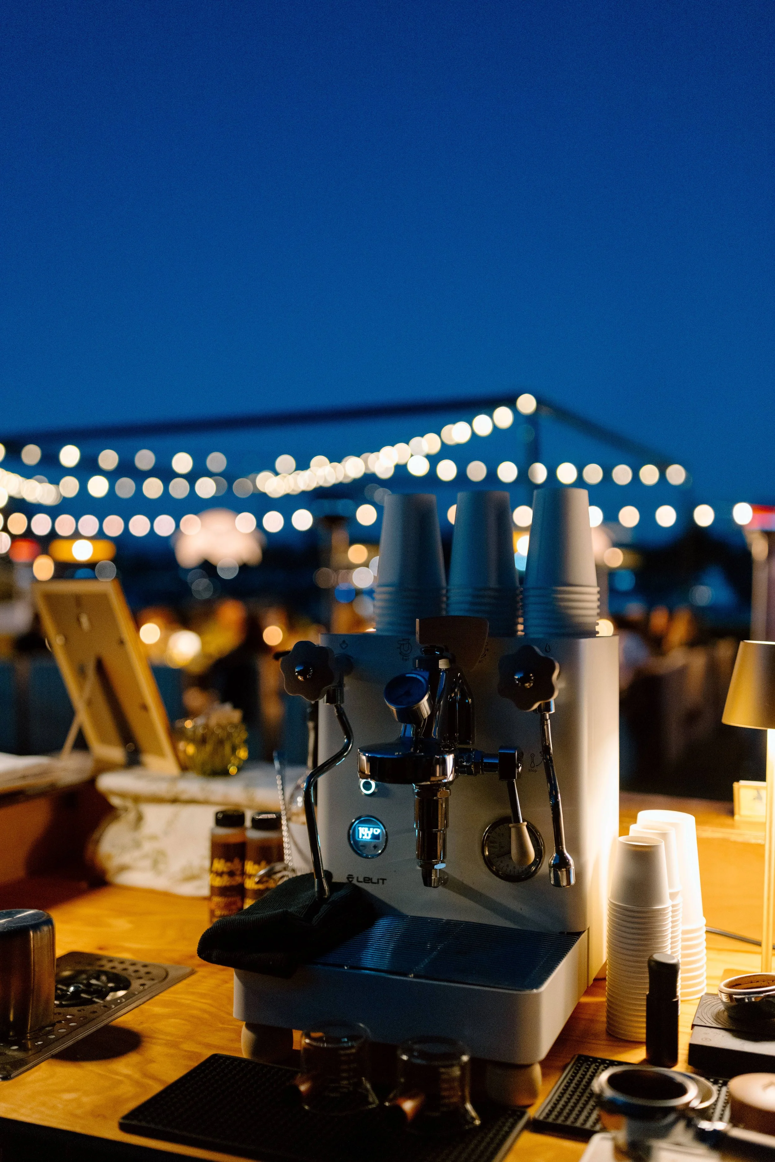 A close-up of a laboratory microscope on a wooden table at an outdoor event during dusk. String lights and blurred background of people and booths are visible.