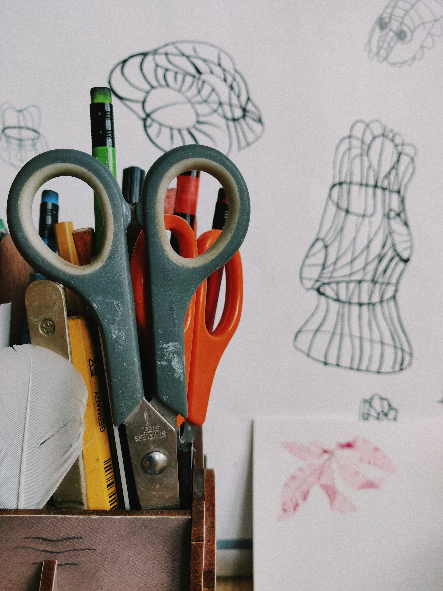A desk organizer holding scissors, pens, and other stationery with a drawing of a wire sculpture of a human head and a print of leaves on a white wall in the background.