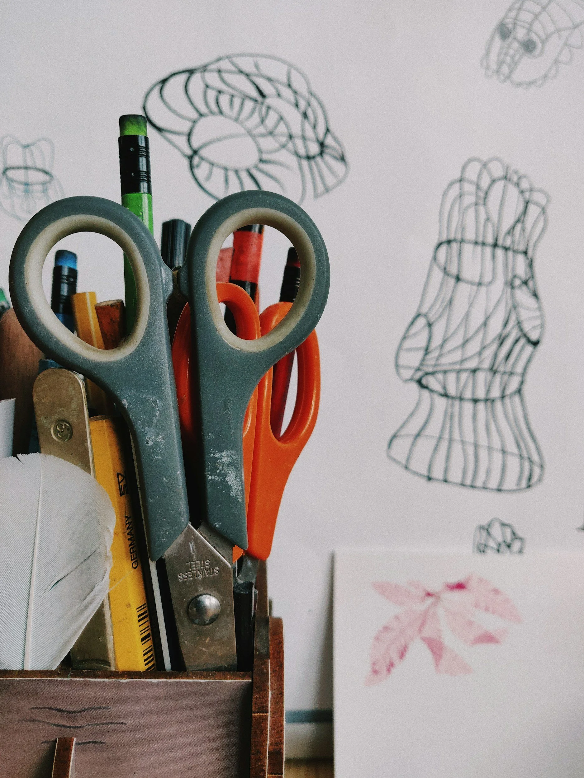 A container holding various scissors, markers, and pens in front of a wall with abstract line art drawings of a head and a leaf.