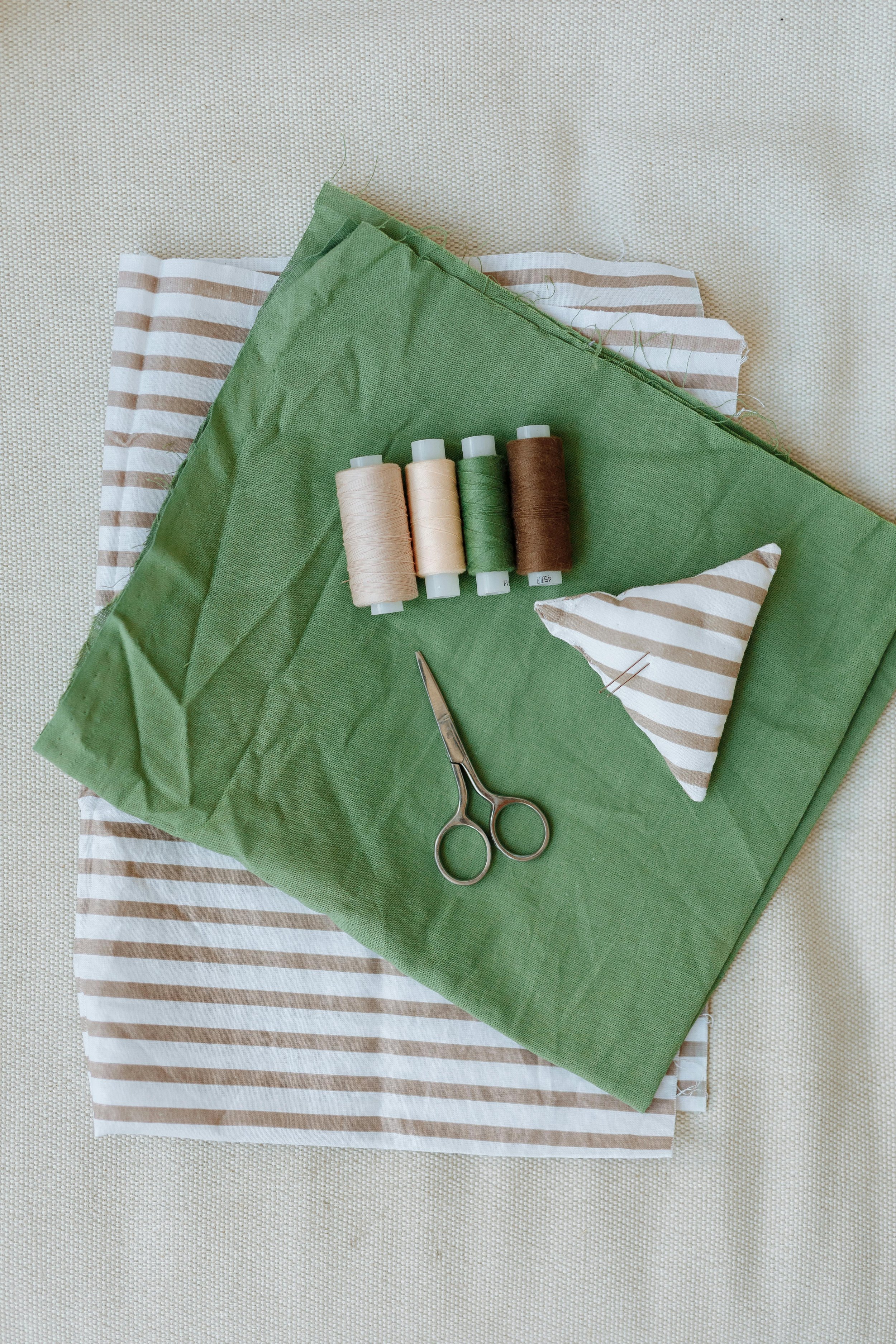 Spools of sewing thread, scissors, and fabric pieces in green and beige with stripes, arranged on a table.