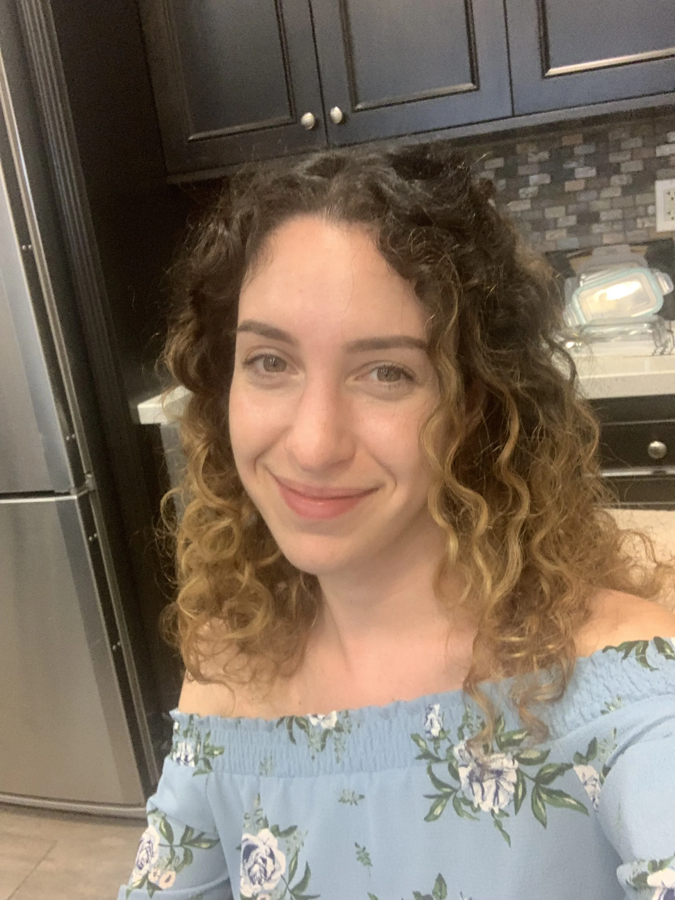 A woman with curly brown hair wearing a light blue off-shoulder dress with a floral pattern, smiling in a kitchen with dark cabinets and a stainless steel appliance.