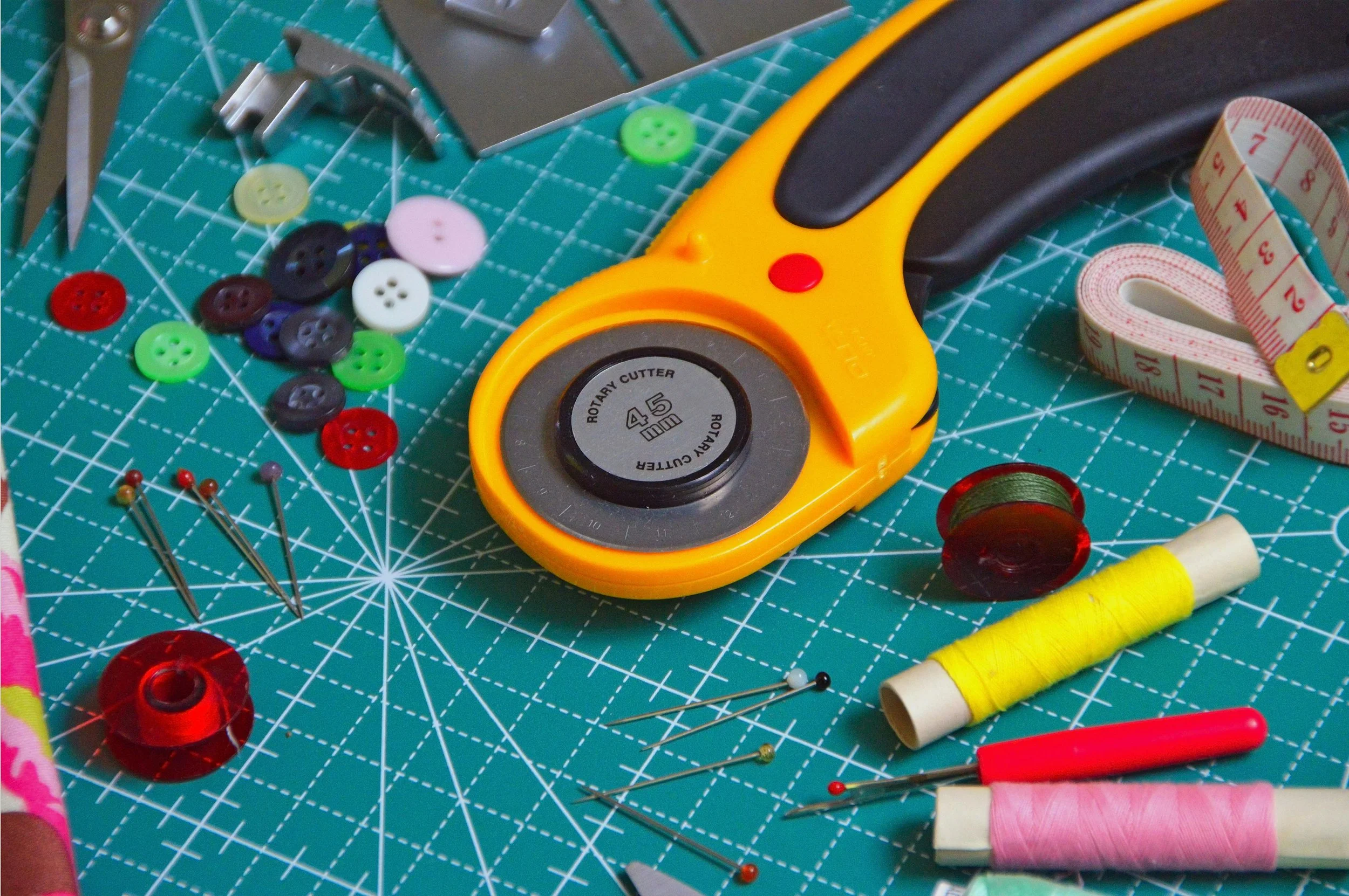 Sewing and crafting tools on a teal cutting mat, including a rotary cutter, buttons in various colors, sewing pins, measuring tape, spools of thread in yellow and pink, and scissors.