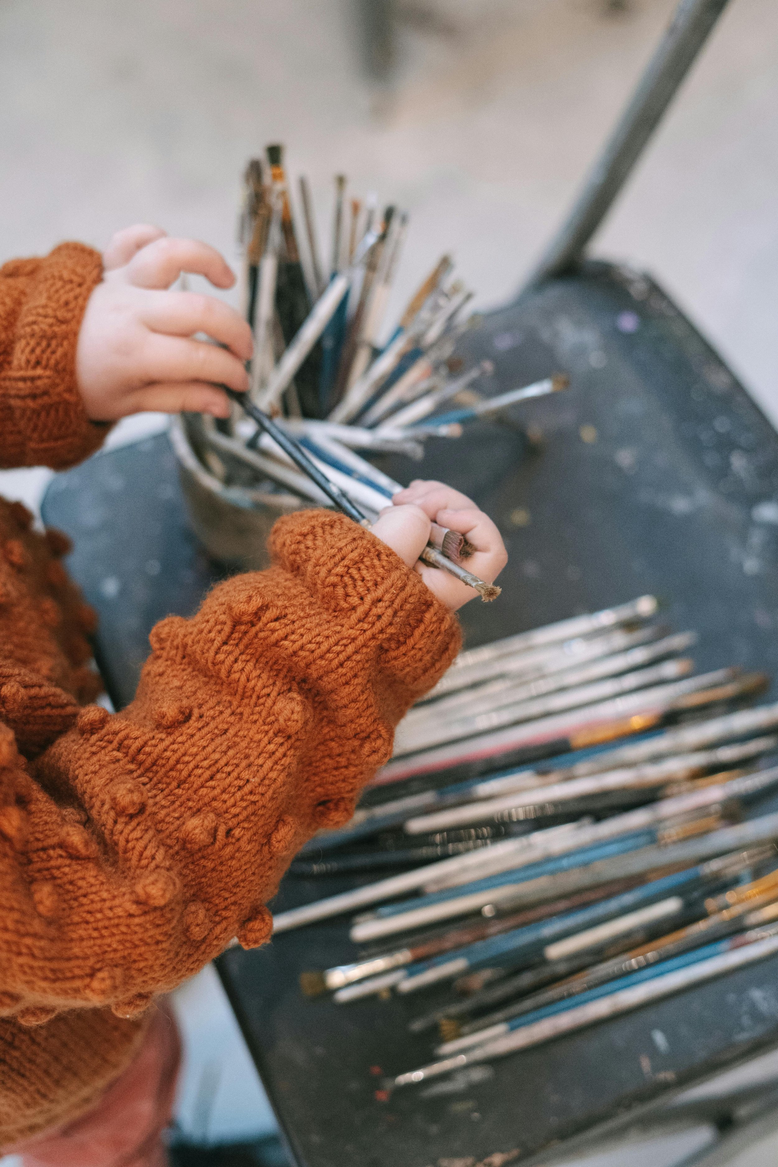 Photo of a child's hands holding paintbrushes, with a container of many brushes on a table.