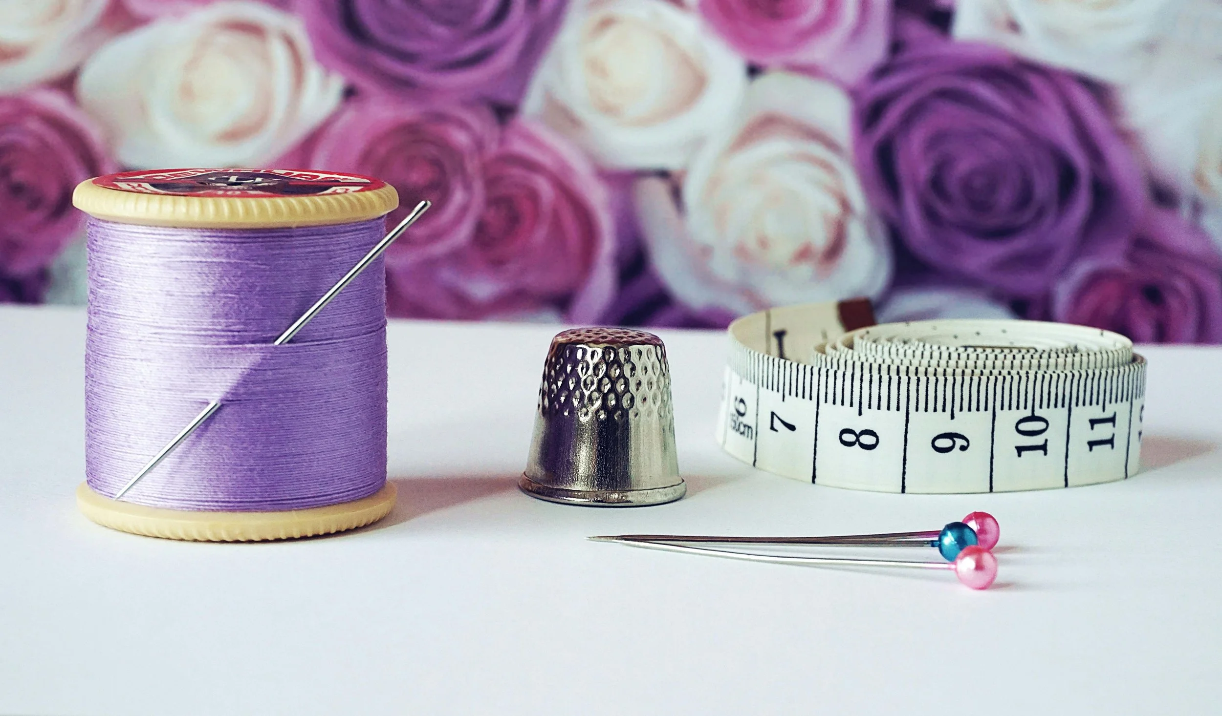 Sewing tools including a spool of purple thread with a needle, a metal thimble, a measuring tape, and two sewing pins with pink and blue heads against a blurred background of purple and white roses.