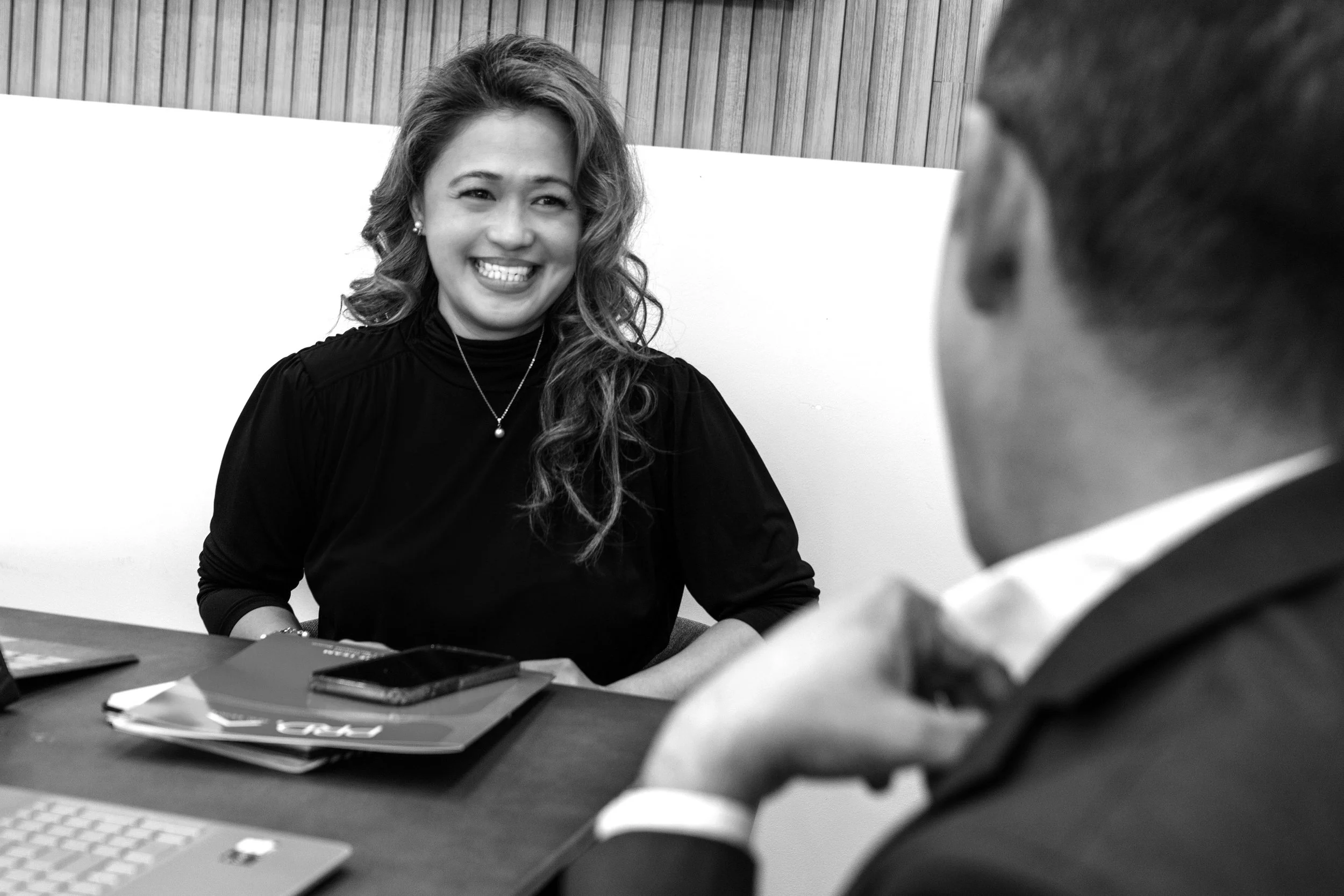 A woman smiling during a conversation with a man in a professional setting, seated at a table with documents and a laptop.