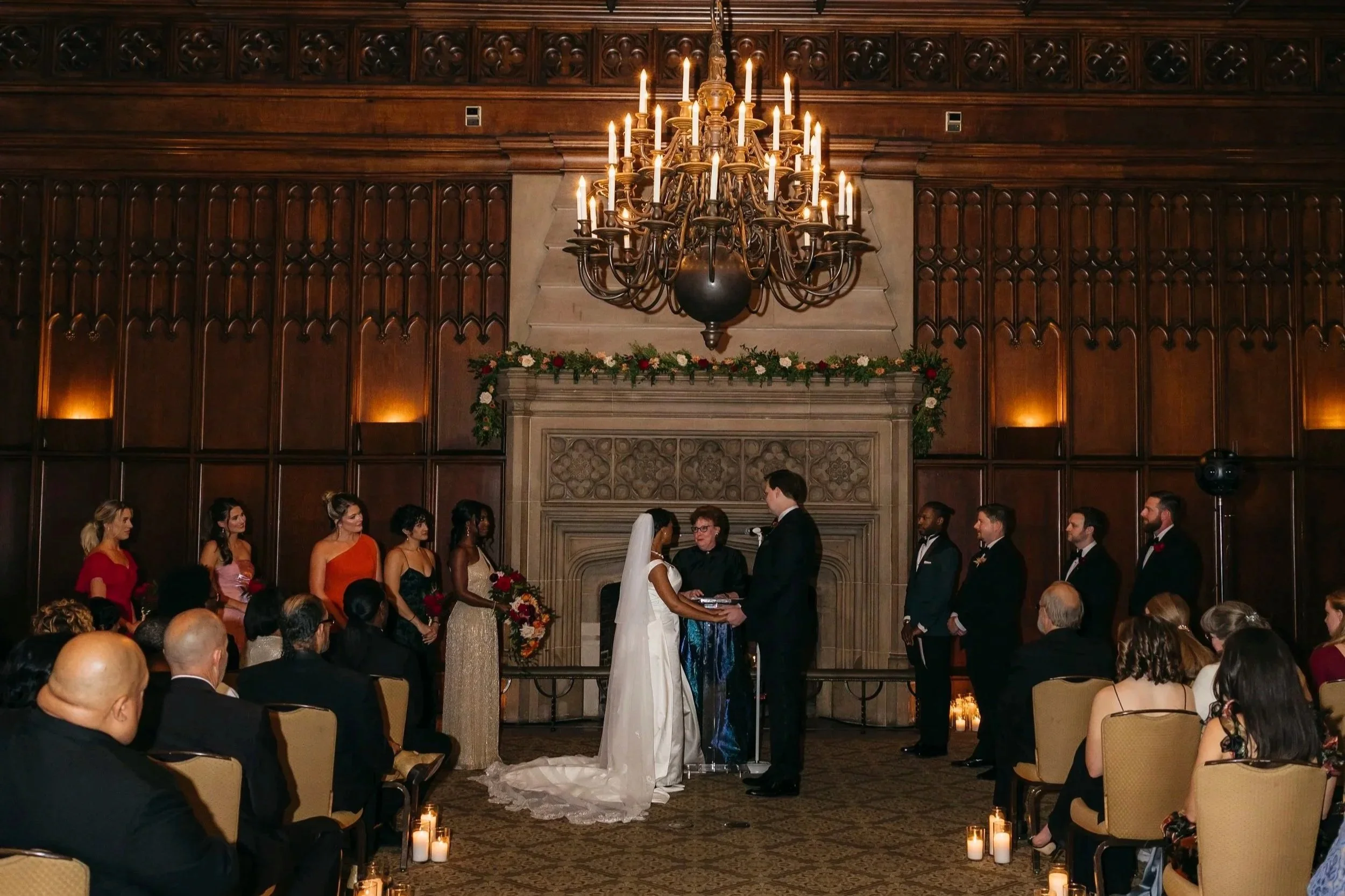 A wedding ceremony taking place in a grand, wood-paneled room with a large chandelier. The bride and groom stand before an officiant, surrounded by bridesmaids and groomsmen. Guests are seated, watching the ceremony.