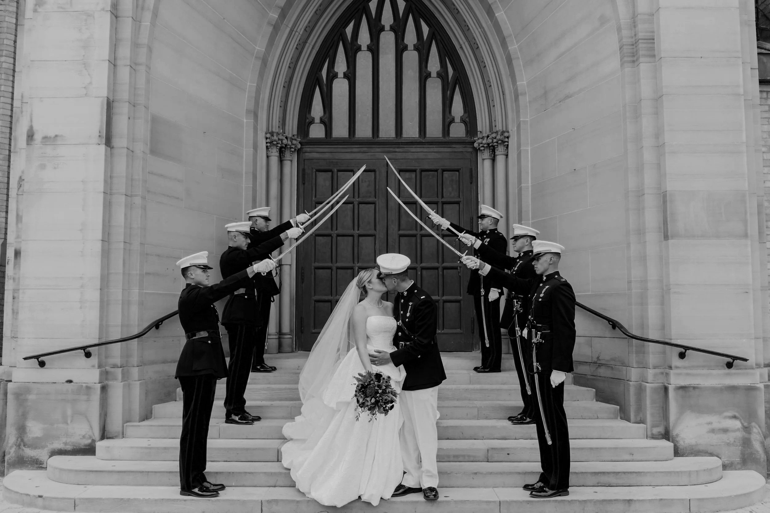 A bride and groom kissing under a Marine Corps sword arch in front of a Cathedral