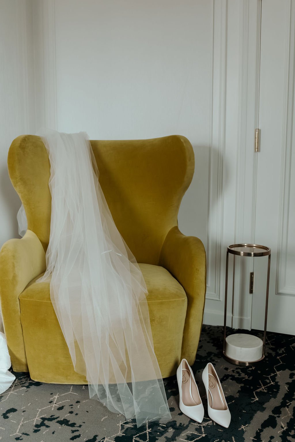 A yellow armchair with a wedding veil draped over one arm and a pair of white high heels on the carpet in front of it while a bride gets ready for her wedding.