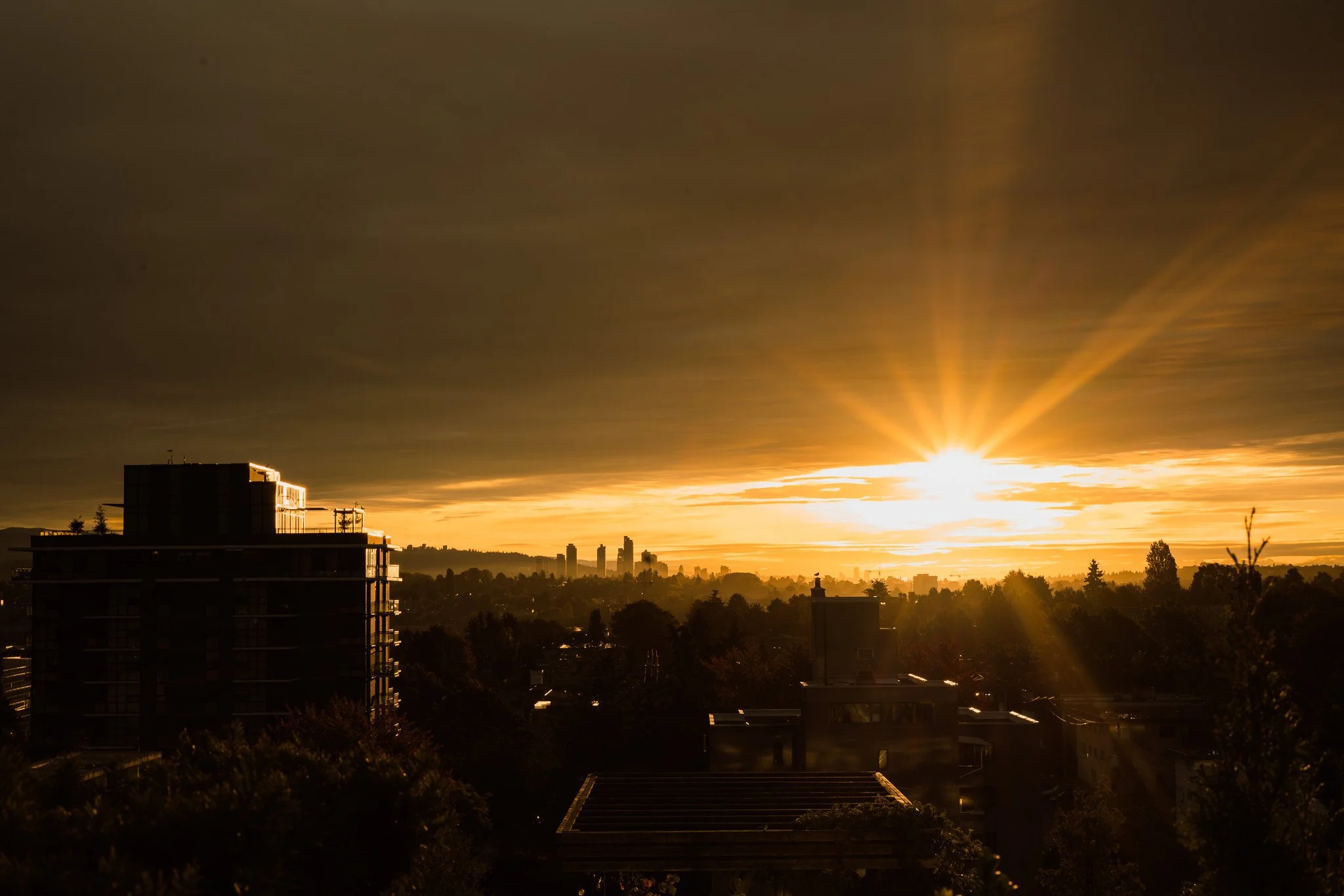 Sunset over a cityscape with buildings silhouetted against a sky with dark clouds and bright, golden sunlight.