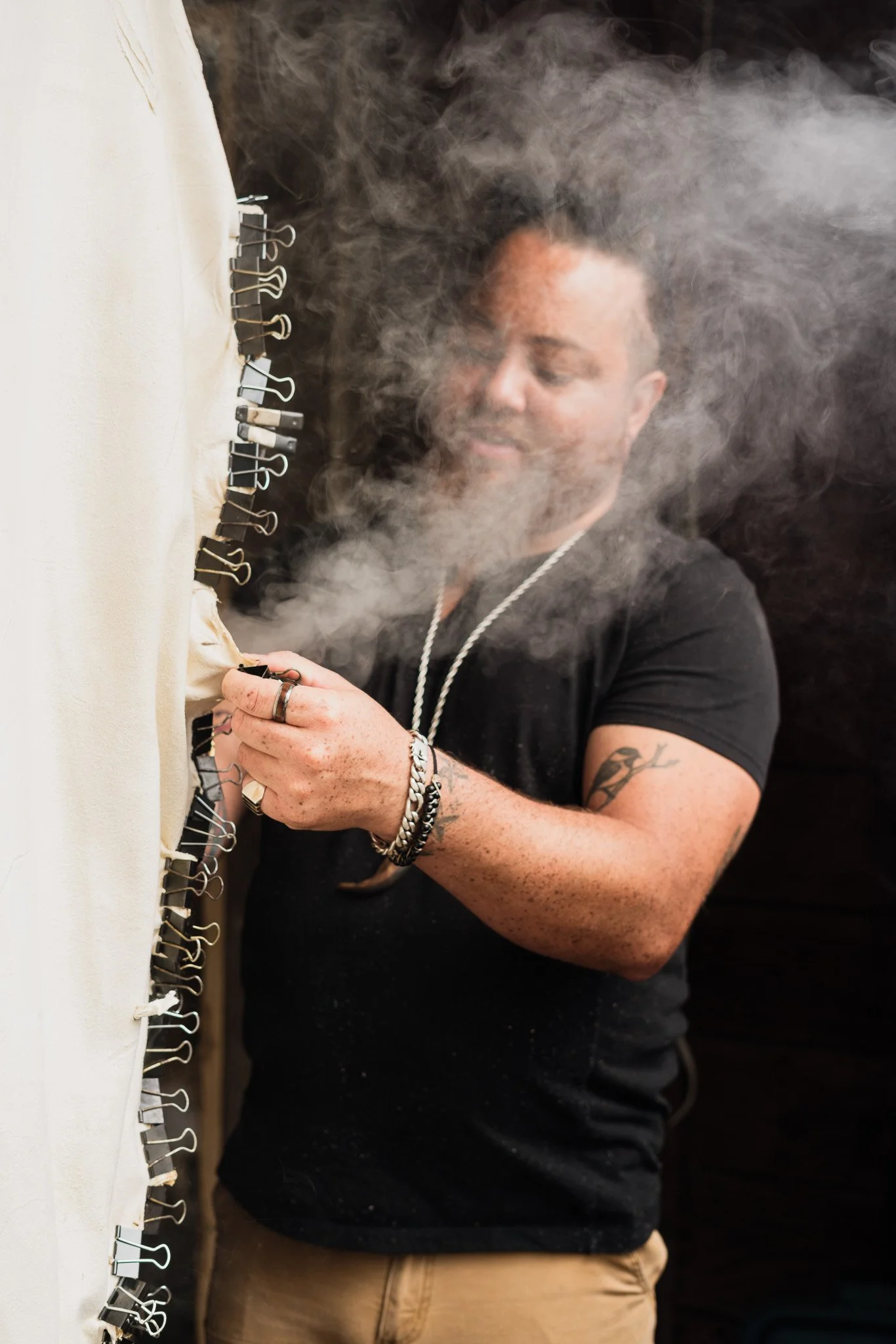 A man with a beard and tattoos on his arm wearing rings, a bracelet, a chain necklace, and a black t-shirt is working with safety pins on a fabric, with steam around him.