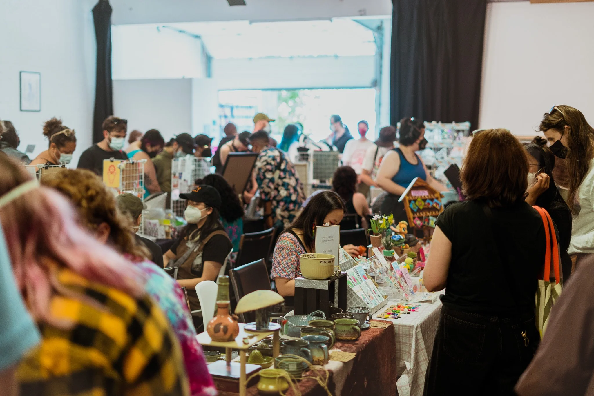 A busy indoor marketplace or craft fair with many people shopping and browsing tables covered with various items for sale, all wearing masks.