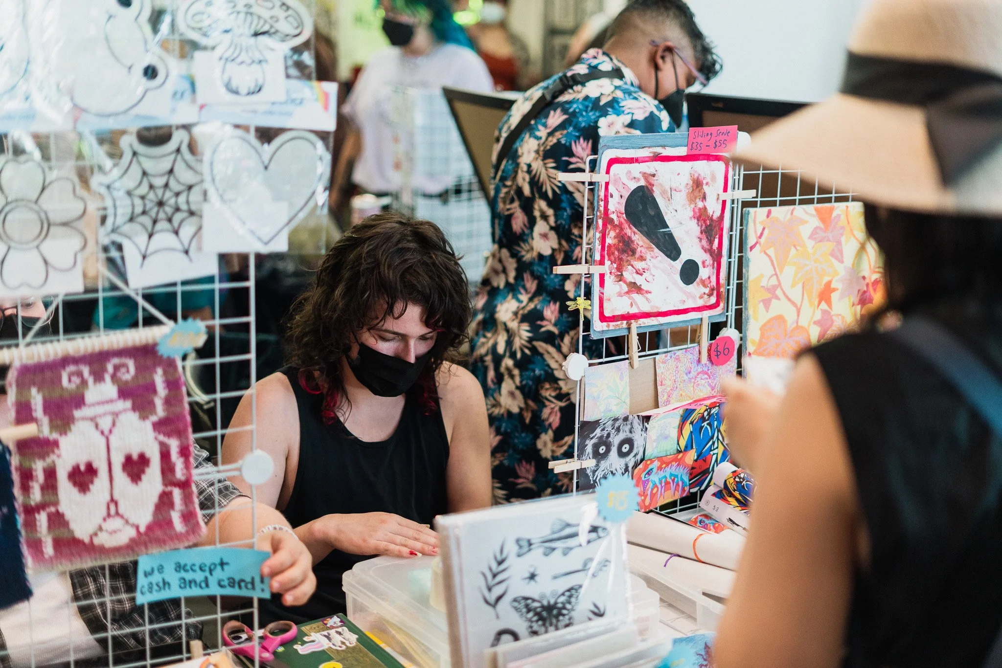 A woman wearing a mask sitting at a booth with various art and craft items, including paper art, stickers, and signs for sale, with other shoppers visible in the background.