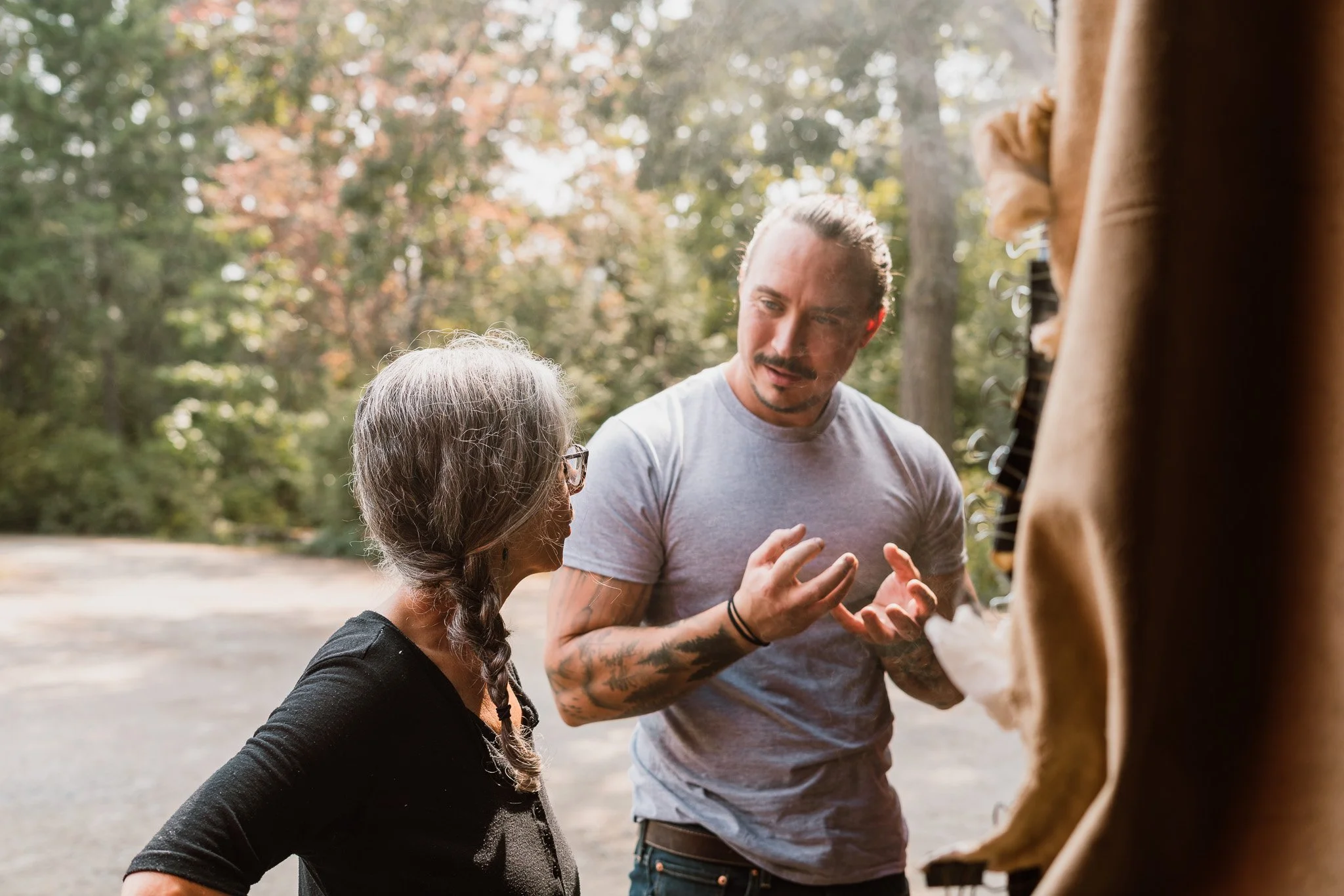 A man is explaining something to an elderly woman outdoors in a forested area while another person, whose face is not visible, stands nearby.