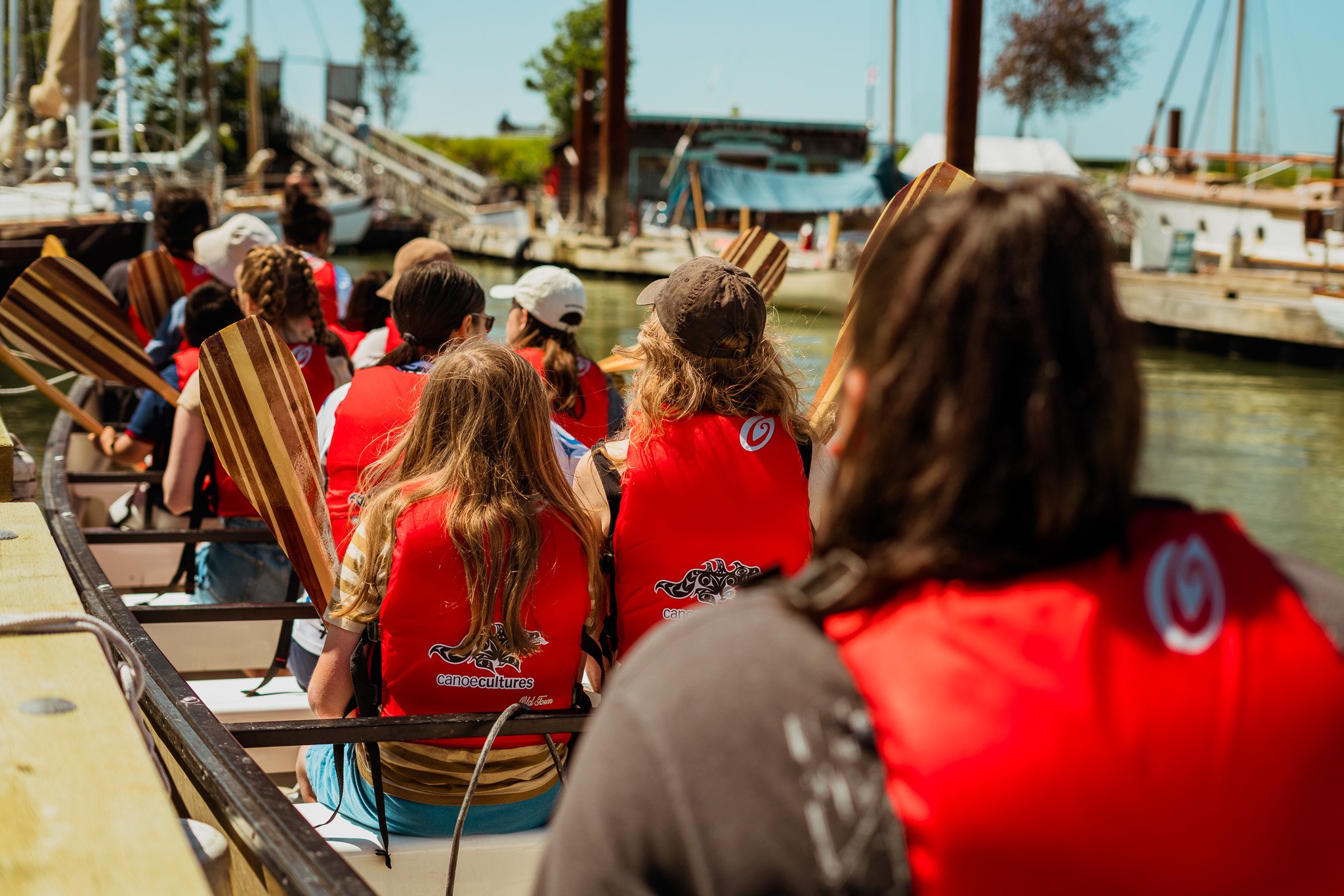 Group of people in red life jackets paddling in a boat on a river or canal, with boats and buildings in the background.