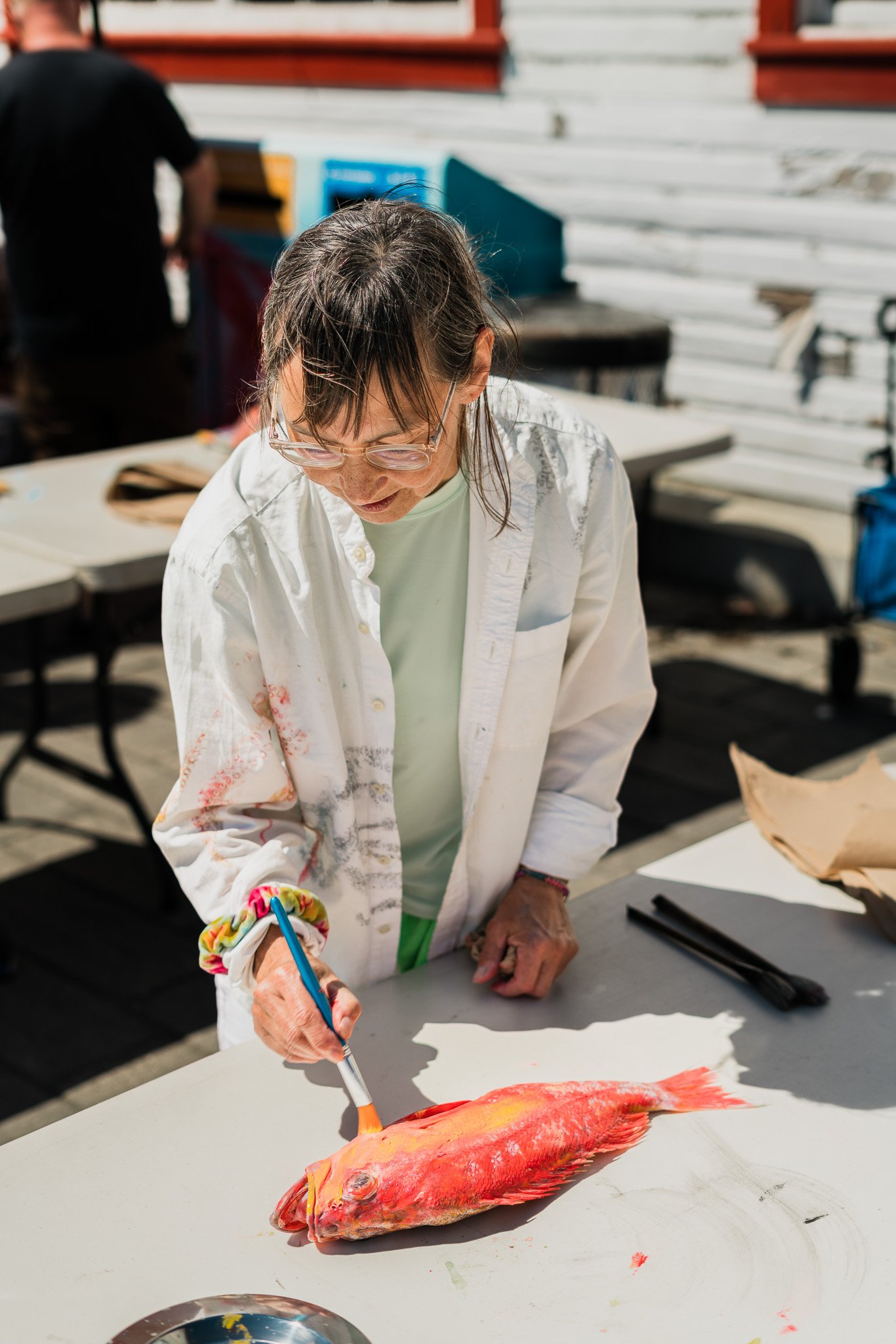 A woman with glasses and a white shirt is painting a red and orange fish on a piece of paper outdoors.
