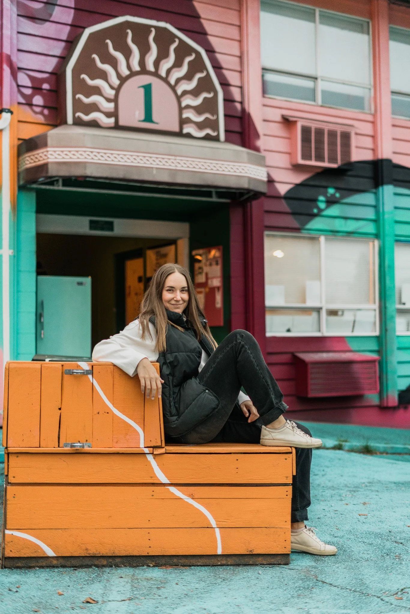 A young woman sitting on an orange wooden bench outdoors in front of colorful, pink and teal buildings with a sign featuring a sun and the number 1. She is smiling, wearing a black vest, white shirt, black pants, and white sneakers.