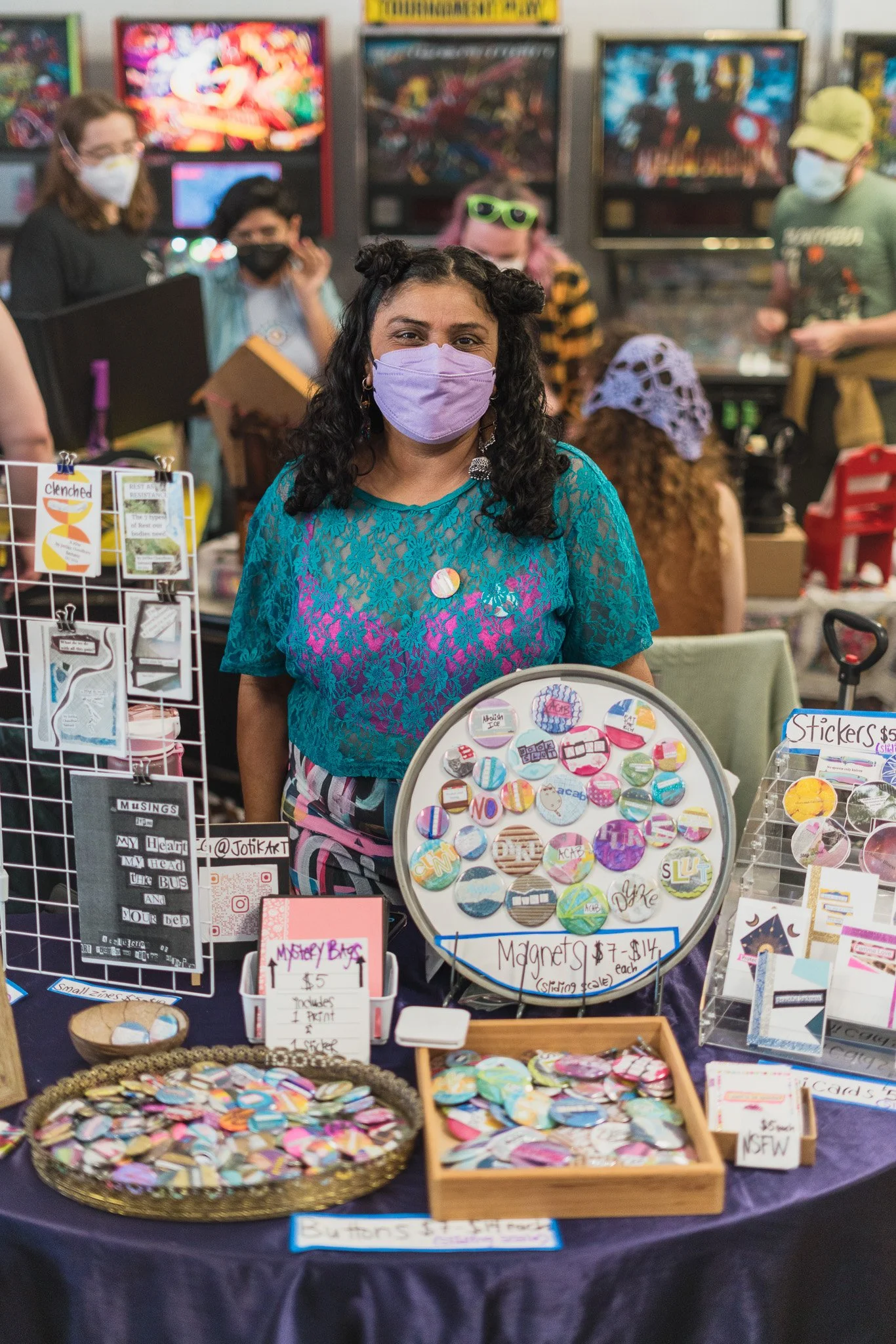 A woman wearing a purple face mask and a teal lace top stands behind a table at a craft fair, displaying colorful button magnets and stickers. Other people with masks are seen in the background, browsing various stalls.