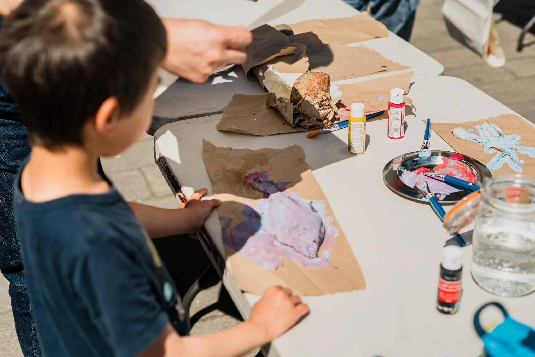 A young boy creating art on brown paper with paintbrushes and various painting supplies on a table.