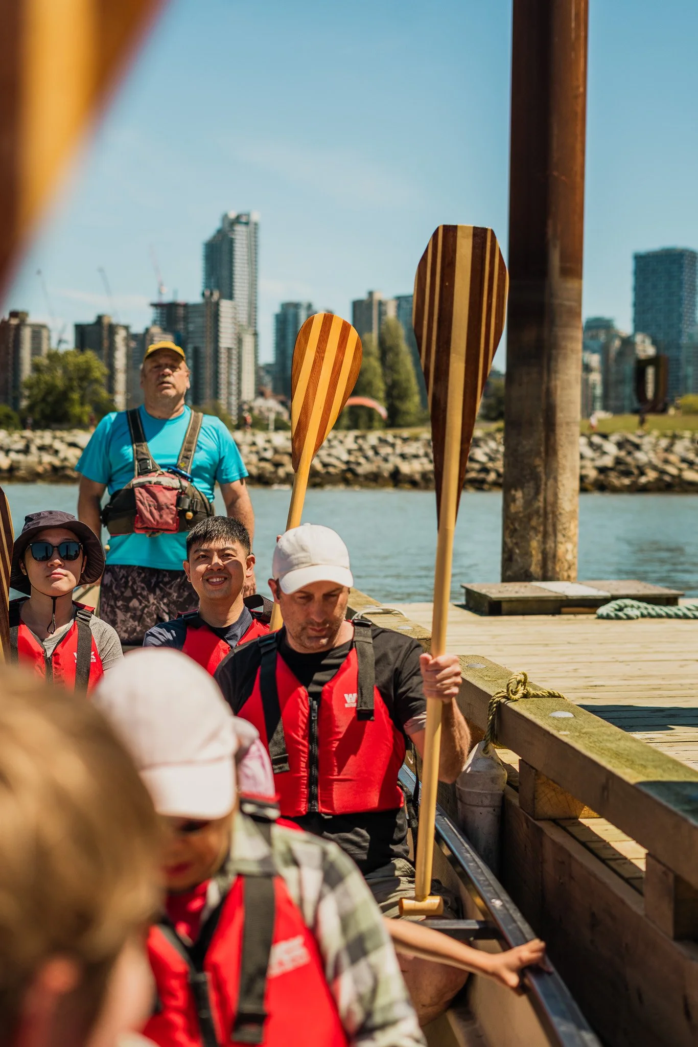 A group of people on a boat holding paddles, preparing to kayak. The scene is set on a sunny day with city buildings in the background.