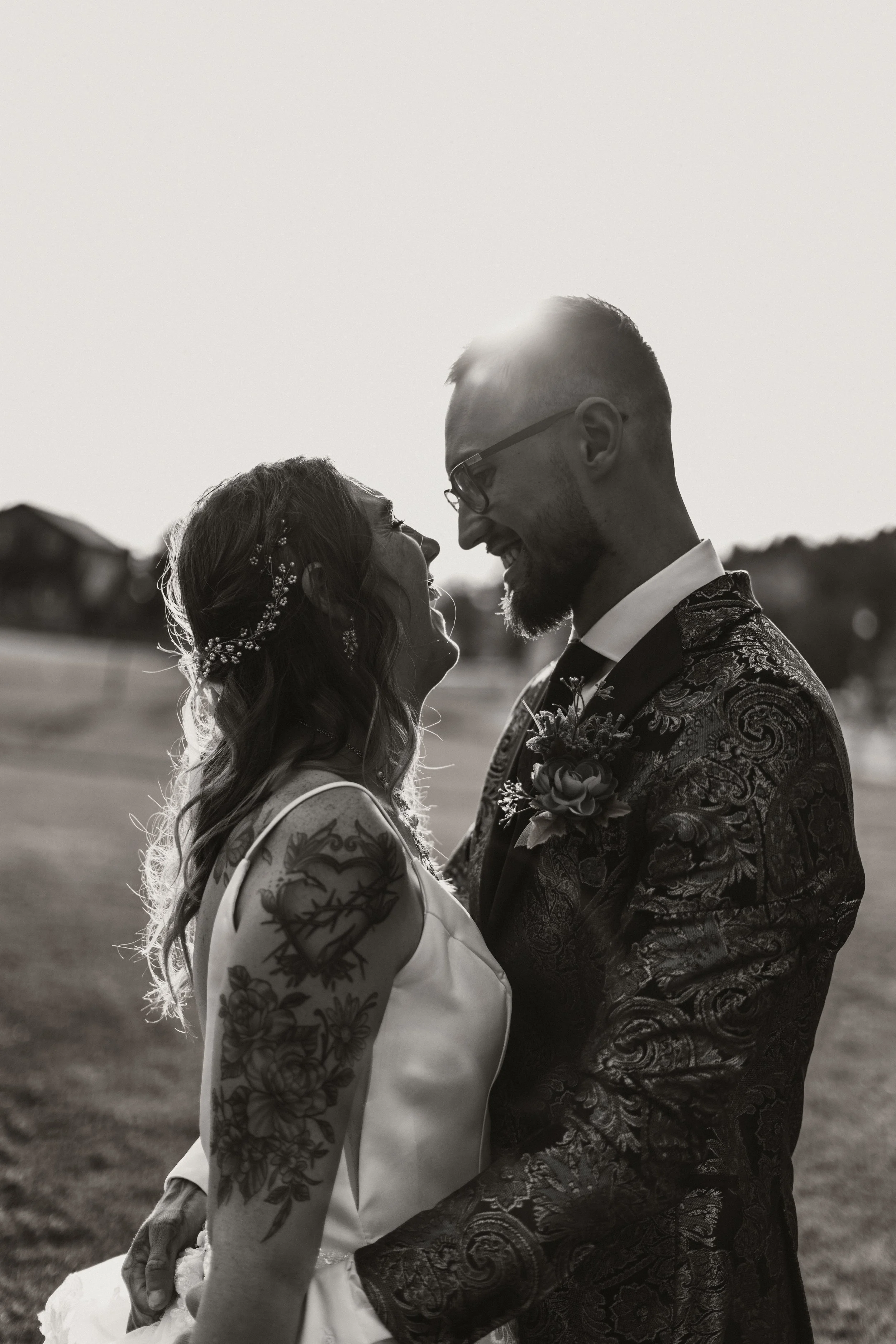 Black and white photo of a couple smiling and looking at each other, standing outdoors during sunset. The woman has tattoos on her arm and is wearing a sleeveless dress with a floral hair accessory. The man is wearing glasses and a patterned suit with a boutonniere. They are holding each other close, sharing a joyful moment.