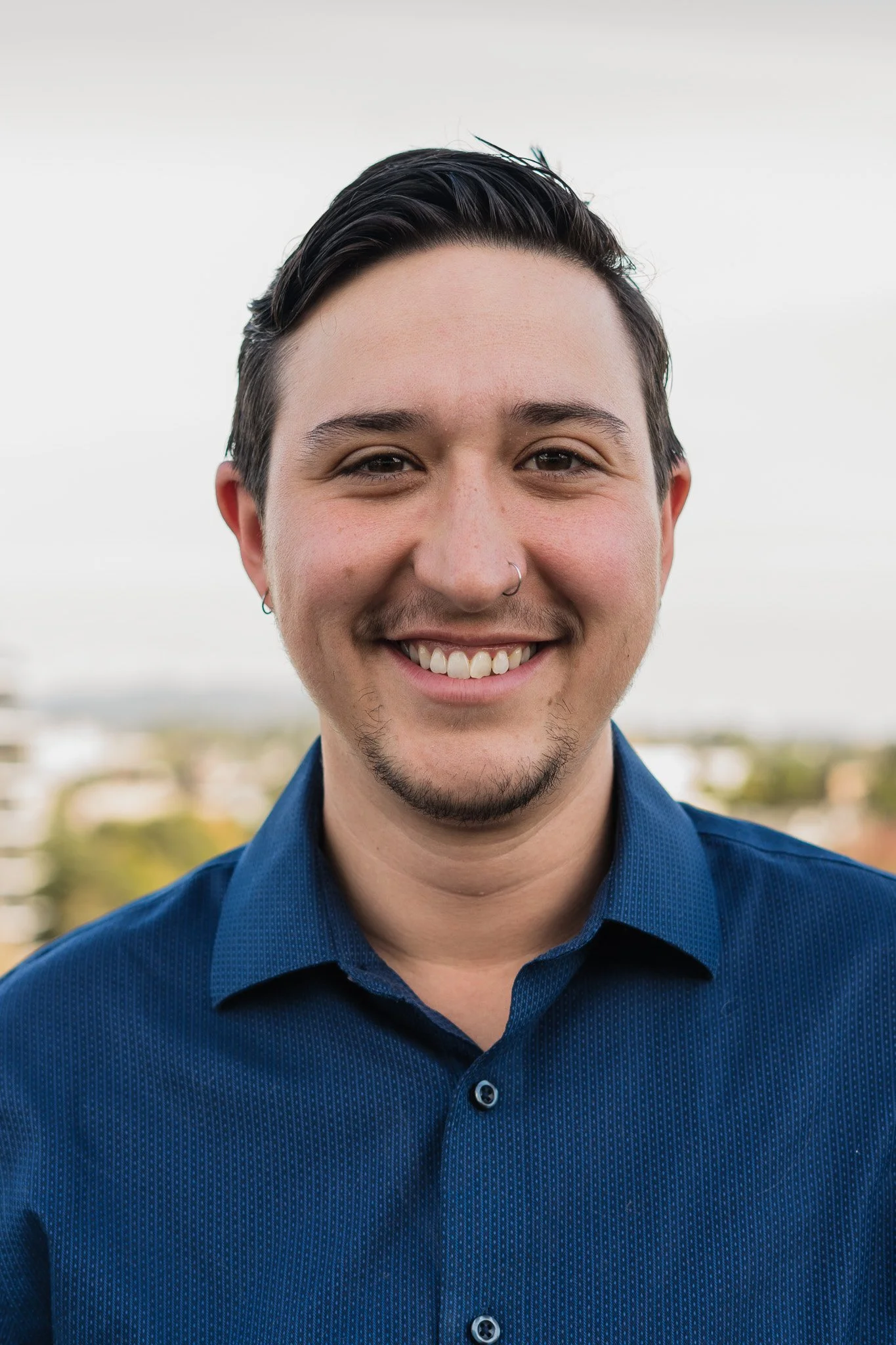 Portrait of a smiling young man with dark, styled hair, light facial hair, piercings, wearing a blue collared shirt, outdoors with a blurred cityscape background.