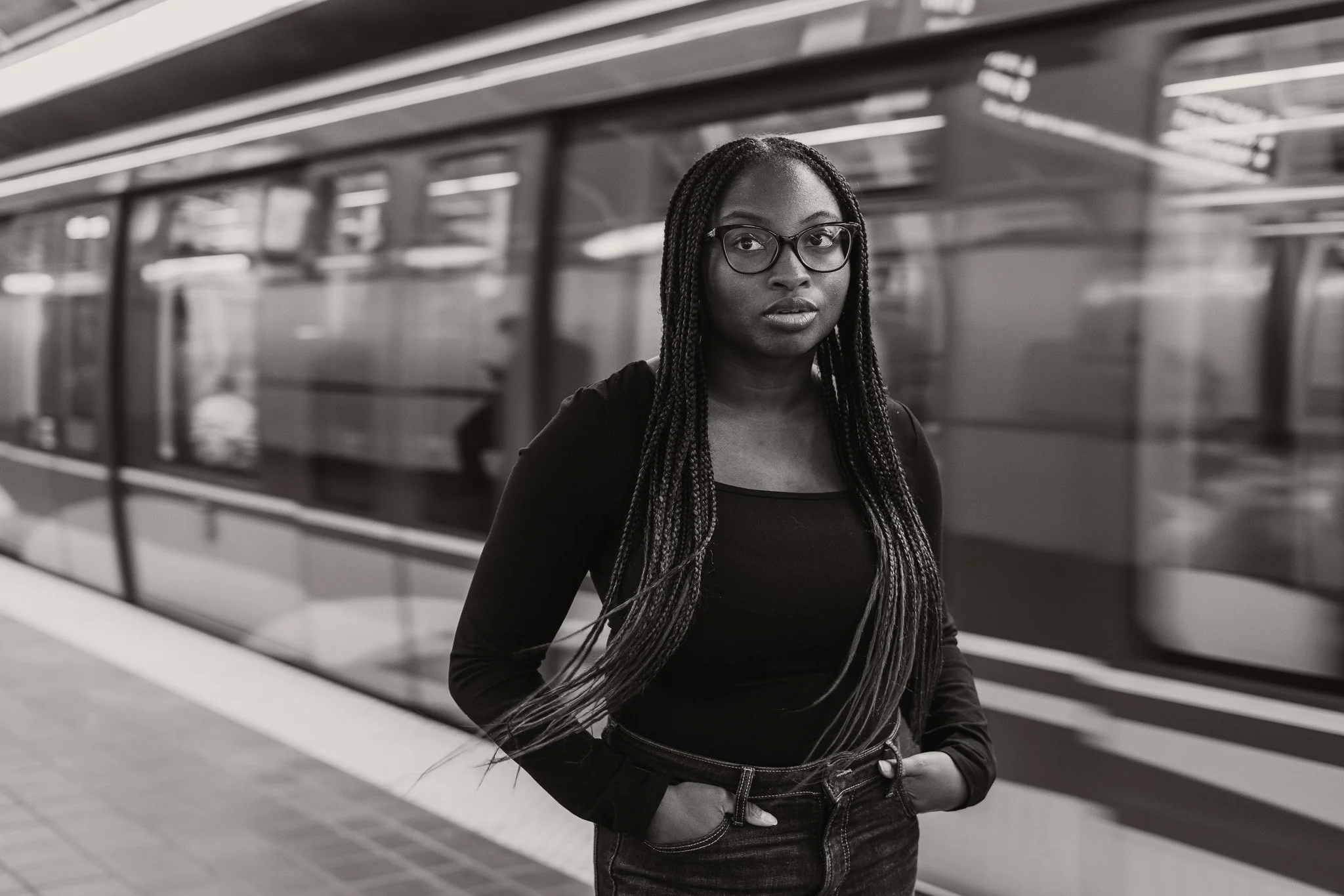 A young woman with braided hair and glasses stands on a train platform with a train in the background.