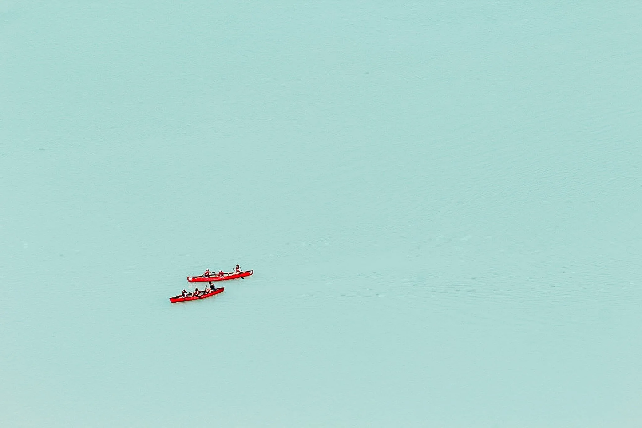 Two red kayaks on a calm, light blue body of water, with a few people paddling in each kayak.