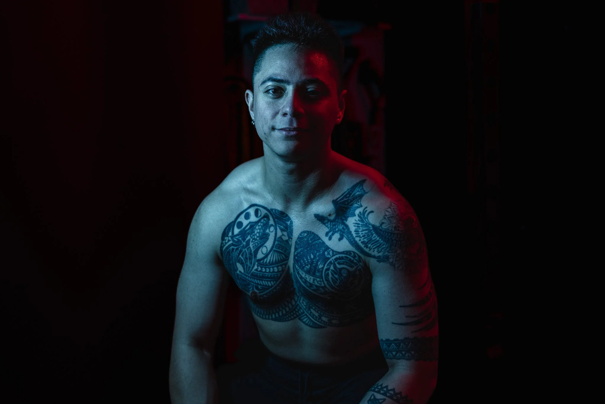 A shirtless man with tattoos on his chest and arm, sitting in a dark room with red and blue lighting.