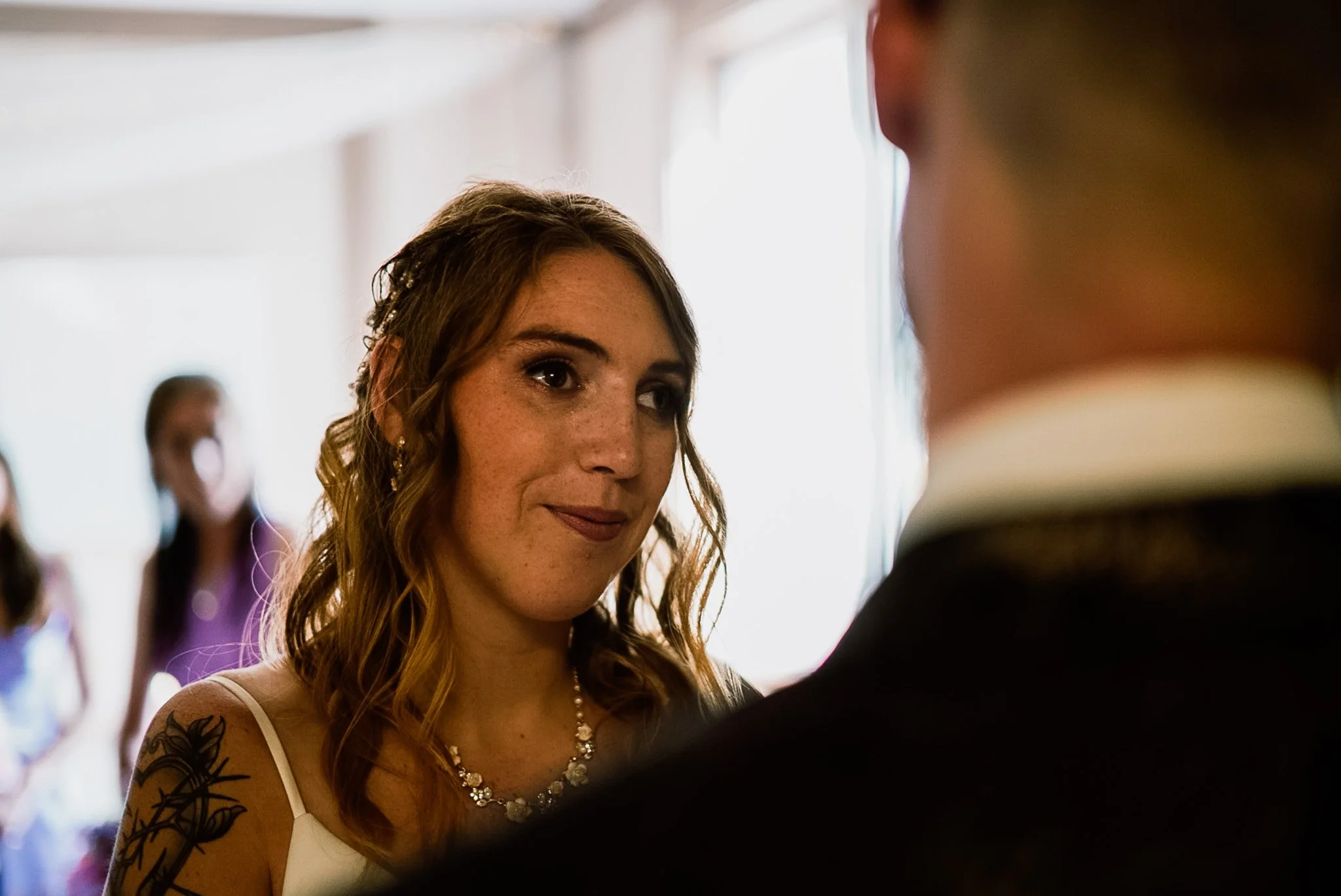 A woman with long wavy hair, freckles, and a necklace, looking at a man during a special event. Other people are blurred in the background.