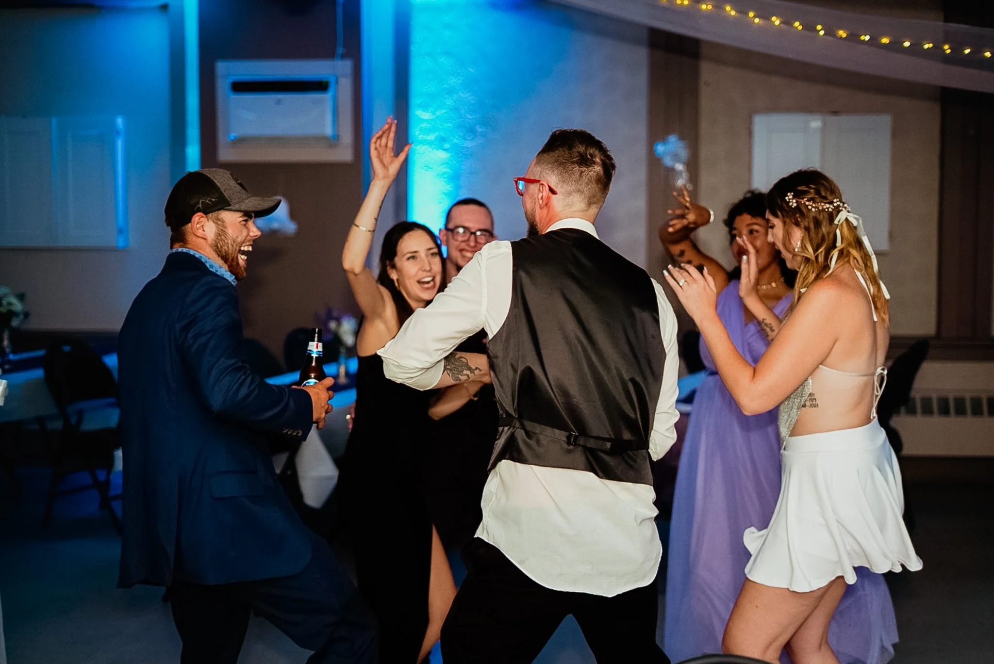 Group of people dancing and celebrating at an indoor party or wedding reception, some holding drinks, under colorful lighting.