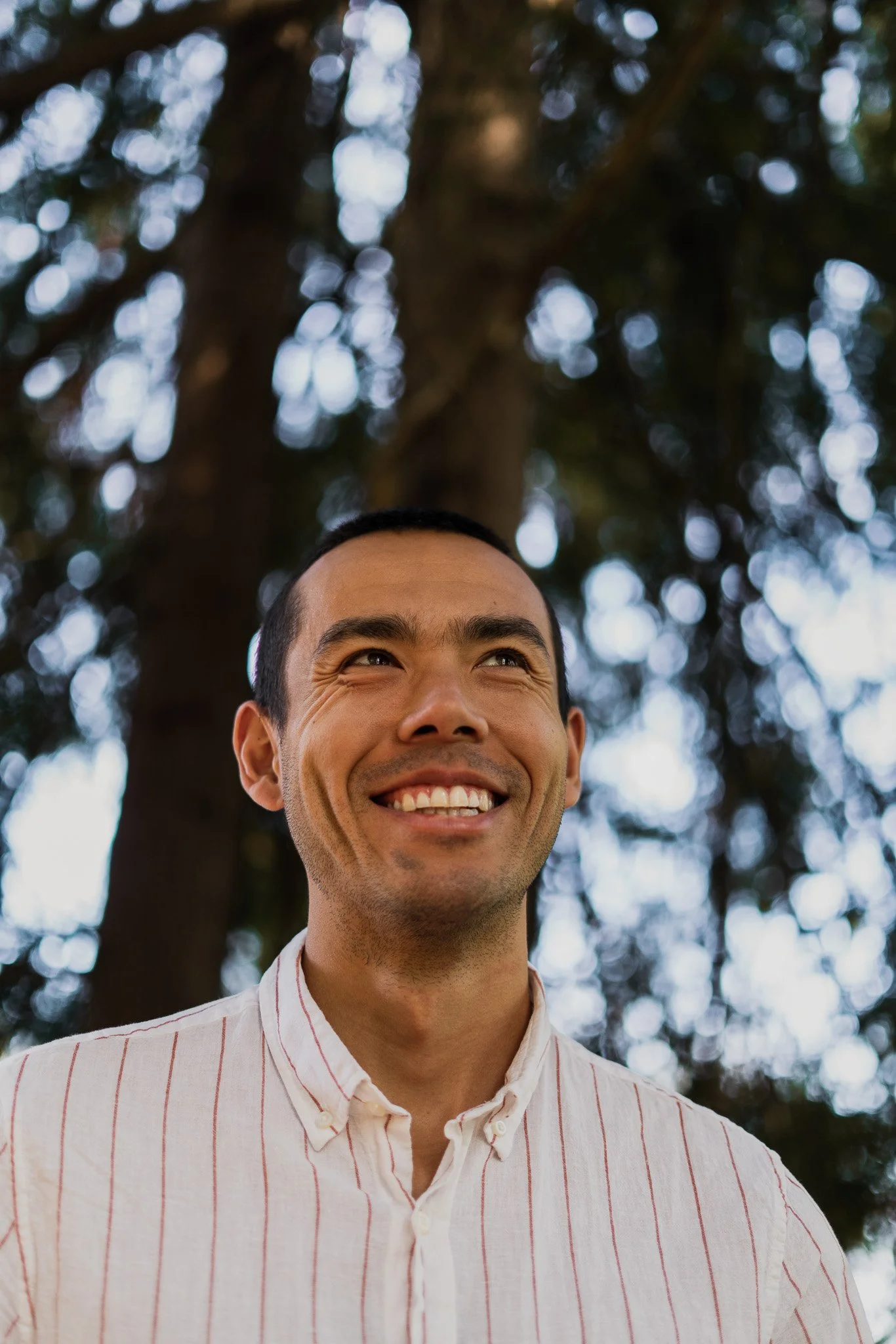Close-up of a smiling man with short dark hair, wearing a white button-up shirt with red pinstripes, standing outdoors with blurred trees and sky in the background.