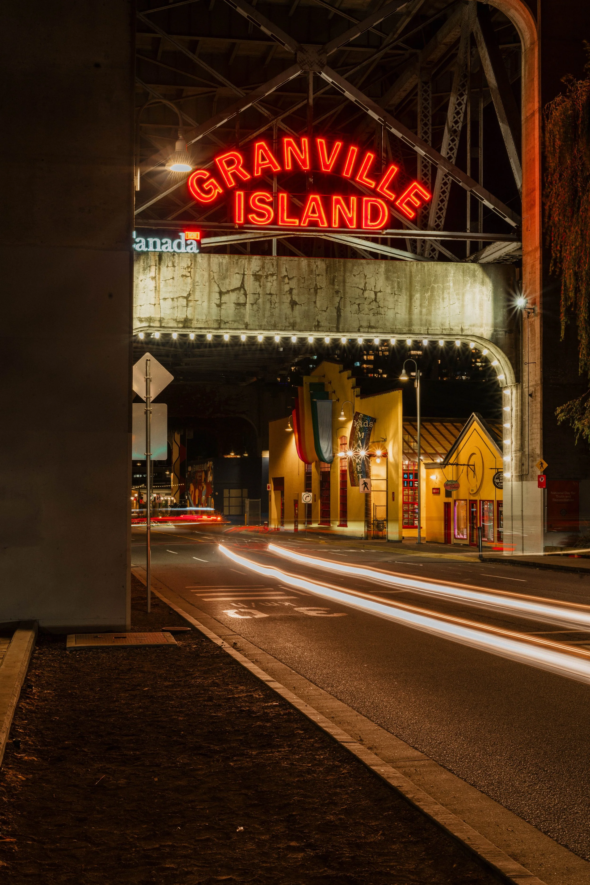 Night view of Granville Island Bridge with neon sign, lit-up shops, colorful flags, and streaks of car lights on the street.