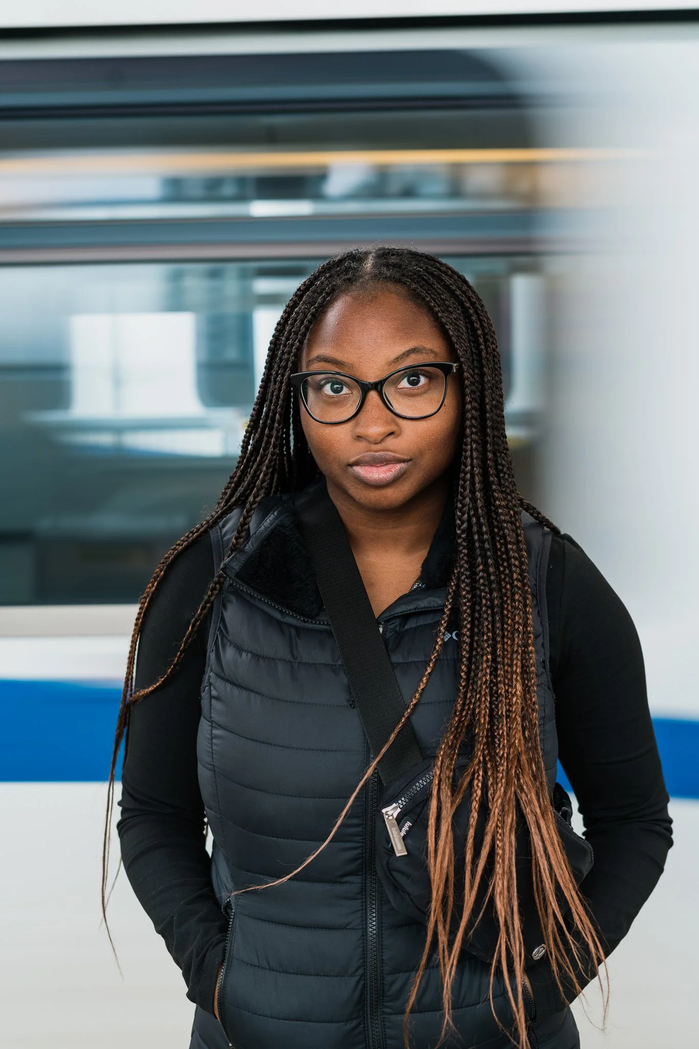 A young woman with long braided hair, glasses, and a black vest standing indoors near a train.