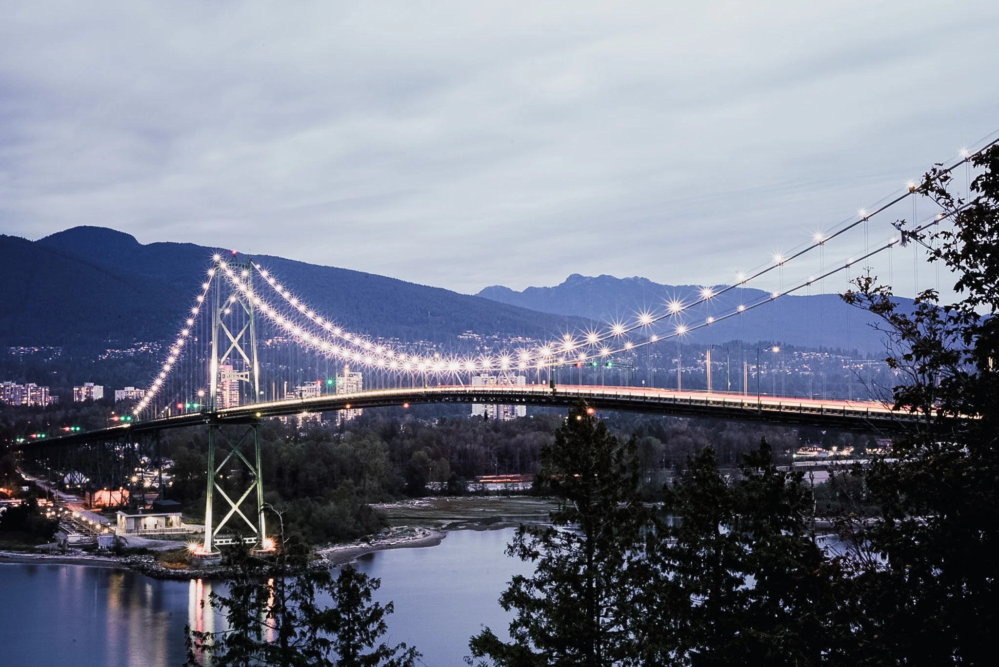 Long exposure photo of a suspension bridge illuminated with lights over a river, with buildings and mountains in the background during dusk.