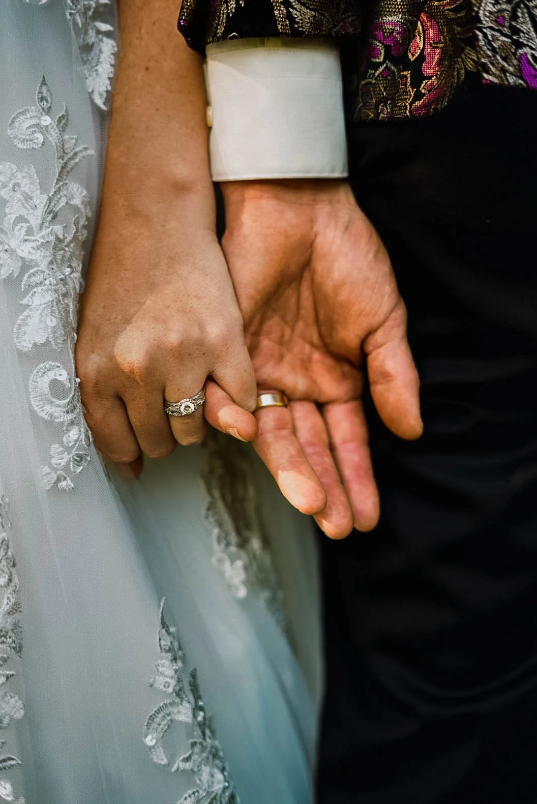 Close-up of a bride and groom holding hands, showing wedding rings. The bride has a lace sleeve and a wedding band with a diamond, and the groom has a plain gold wedding band.