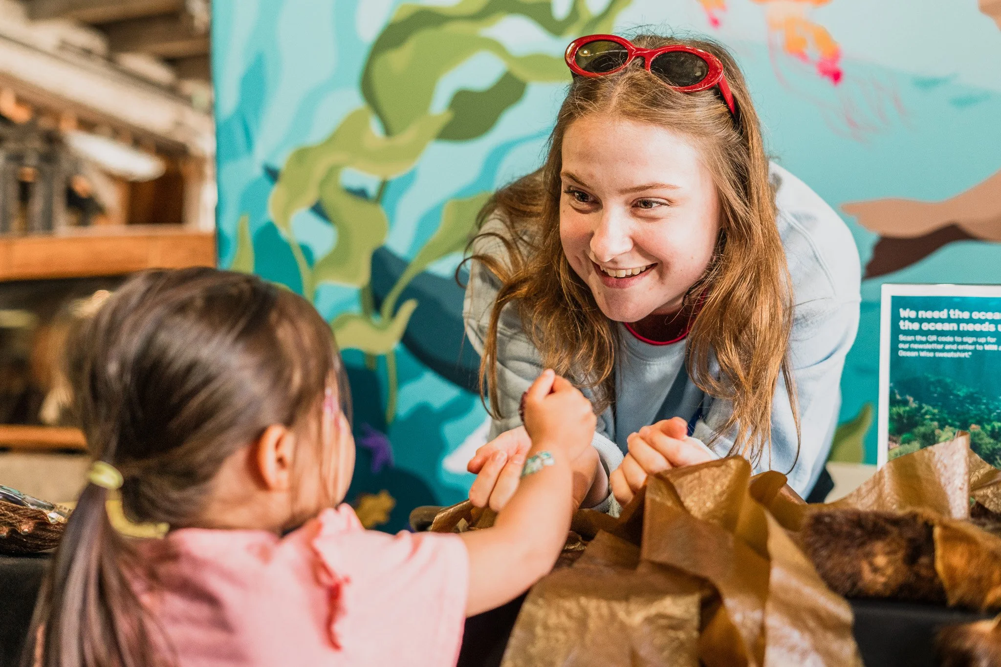 A young woman with red hair, wearing sunglasses on her head, smiling and engaging with a child with brown hair pulled back in a ponytail. They are arm-wrestling at an event with an ocean-themed mural in the background.