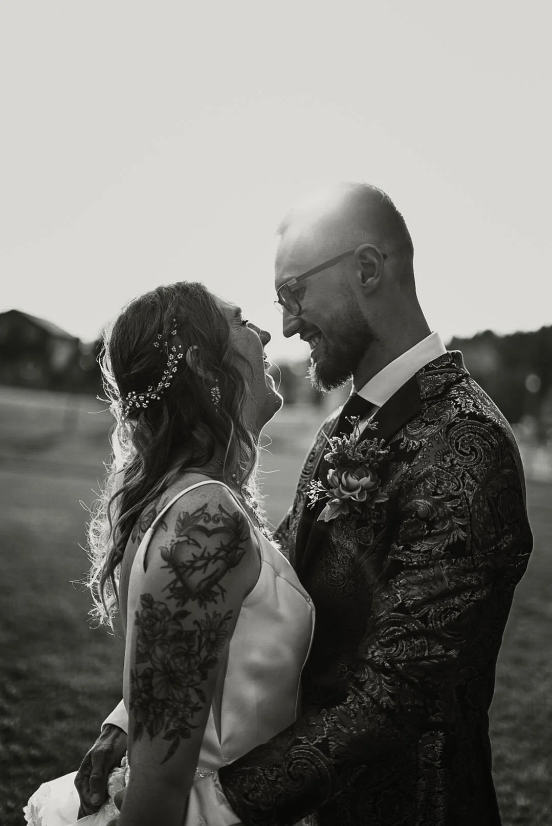 A black-and-white photo of a bride and groom smiling at each other outdoors during sunset, with the sun behind them. The bride has long, wavy hair with floral accessories and visible tattoos on her arm. The groom wears glasses and a patterned suit.