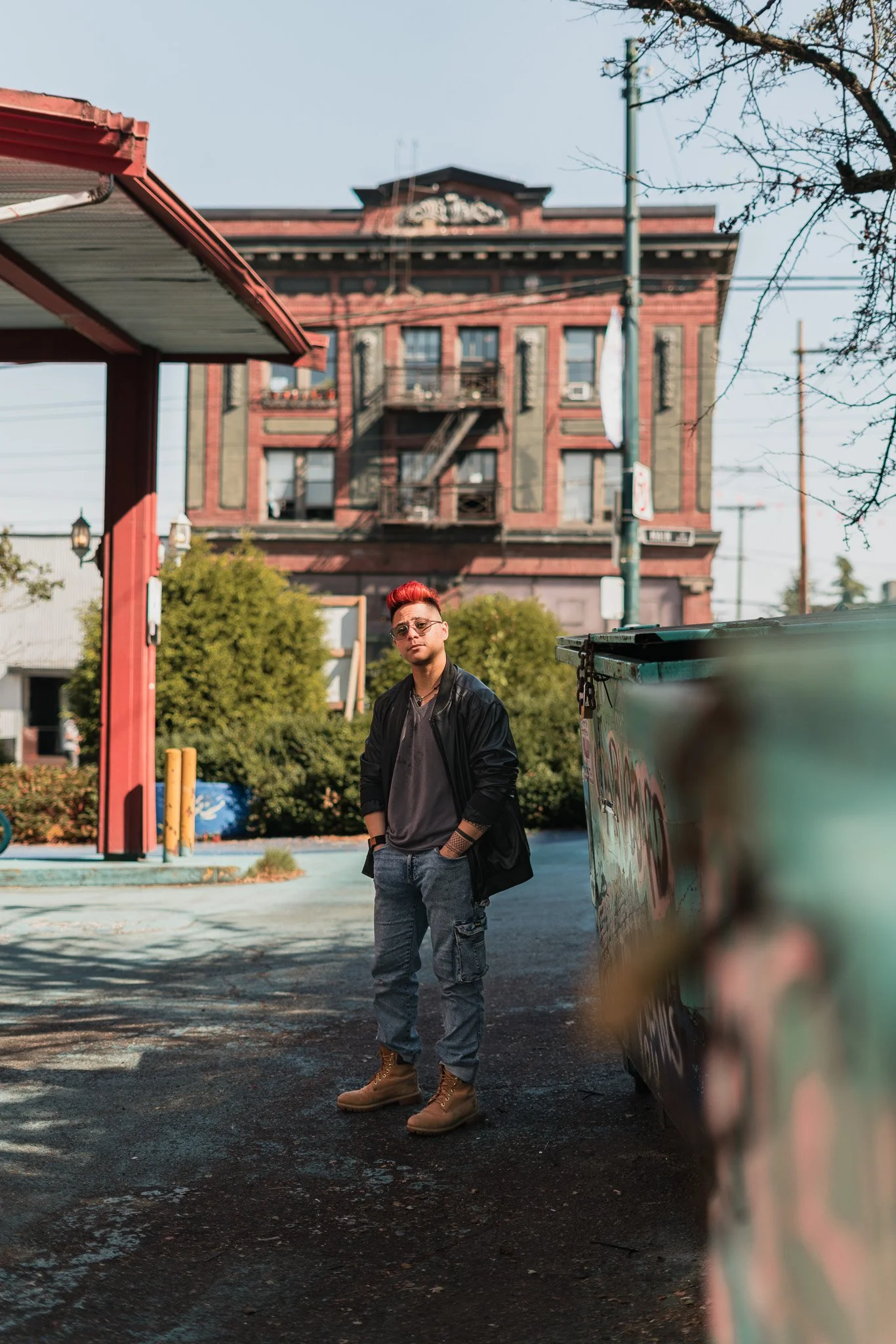 A young man with red hair and glasses standing near a graffiti-covered dumpster outside on an asphalt lot, with a historic red brick building in the background.