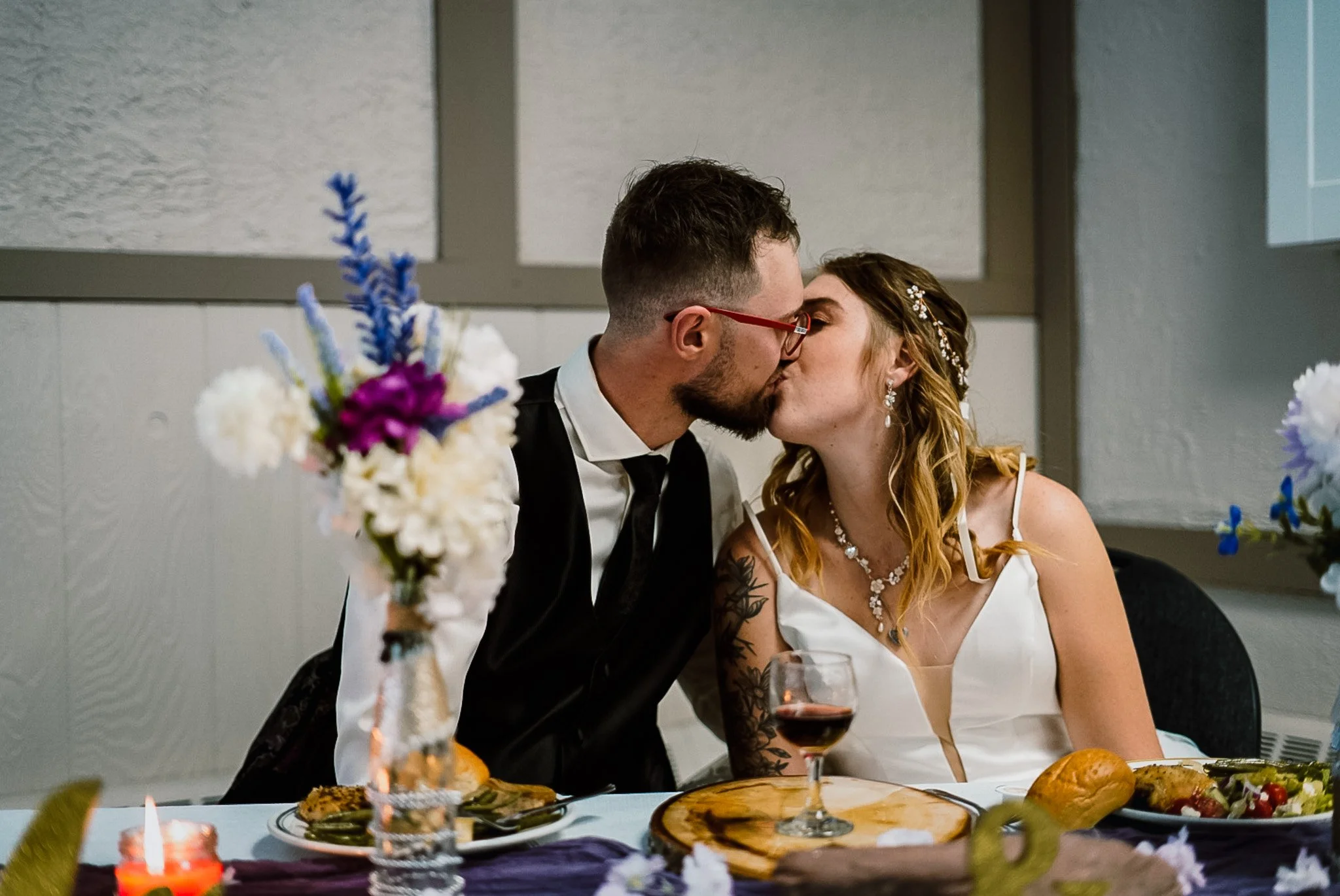 A man and woman sharing a kiss at a wedding reception, seated at a decorated table with flowers, bread, wine, and other foods.