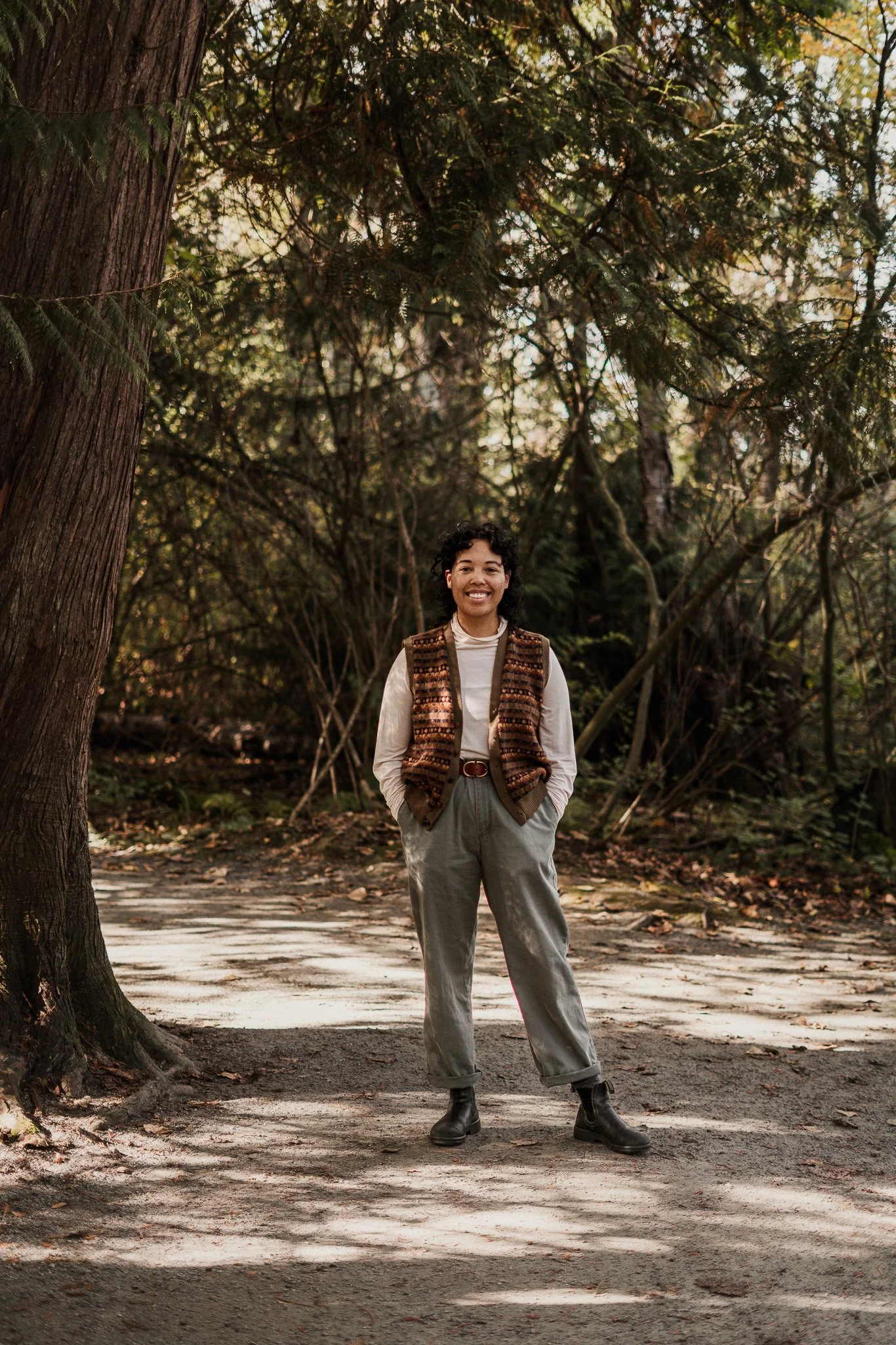 A woman standing outdoors on a dirt path in a forest, smiling with her hands in her pockets, wearing a beige sweater, patterned vest, gray pants, and black boots.