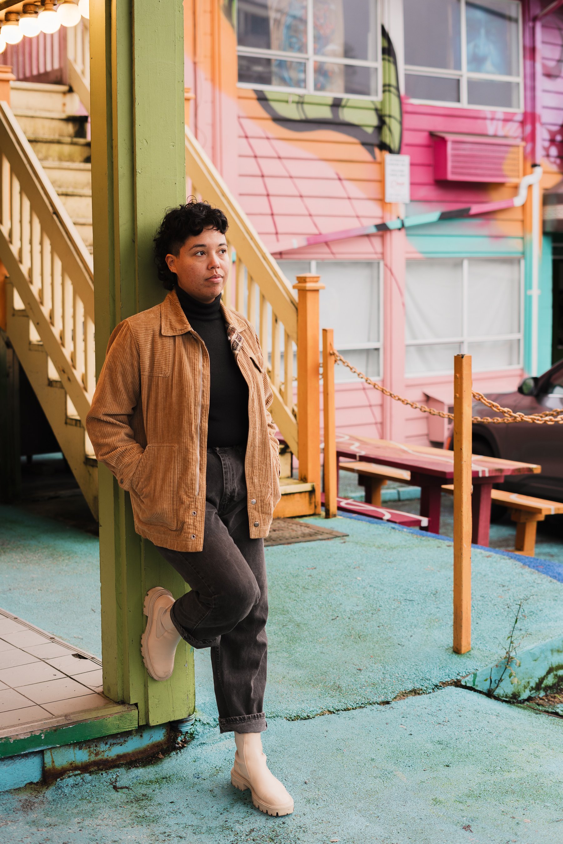 A person leaning against a green post outside a colorful building with pink, orange, and teal walls. The person wears a brown jacket, black turtleneck, gray jeans, and white boots, with their hands in their pockets.