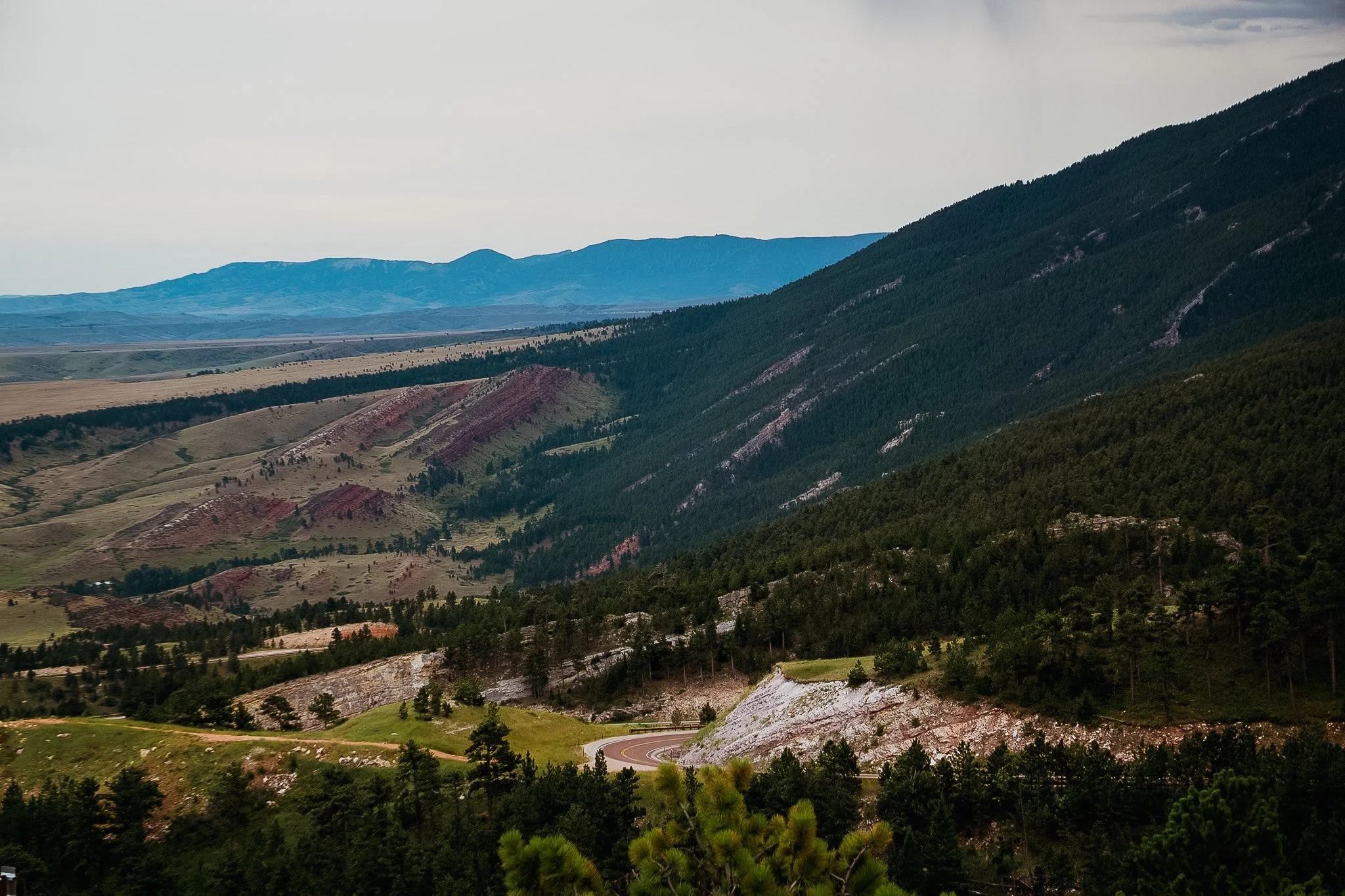 A mountainous landscape with green forested slopes, red and white rock formations, and distant mountains in the background under a cloudy sky.
