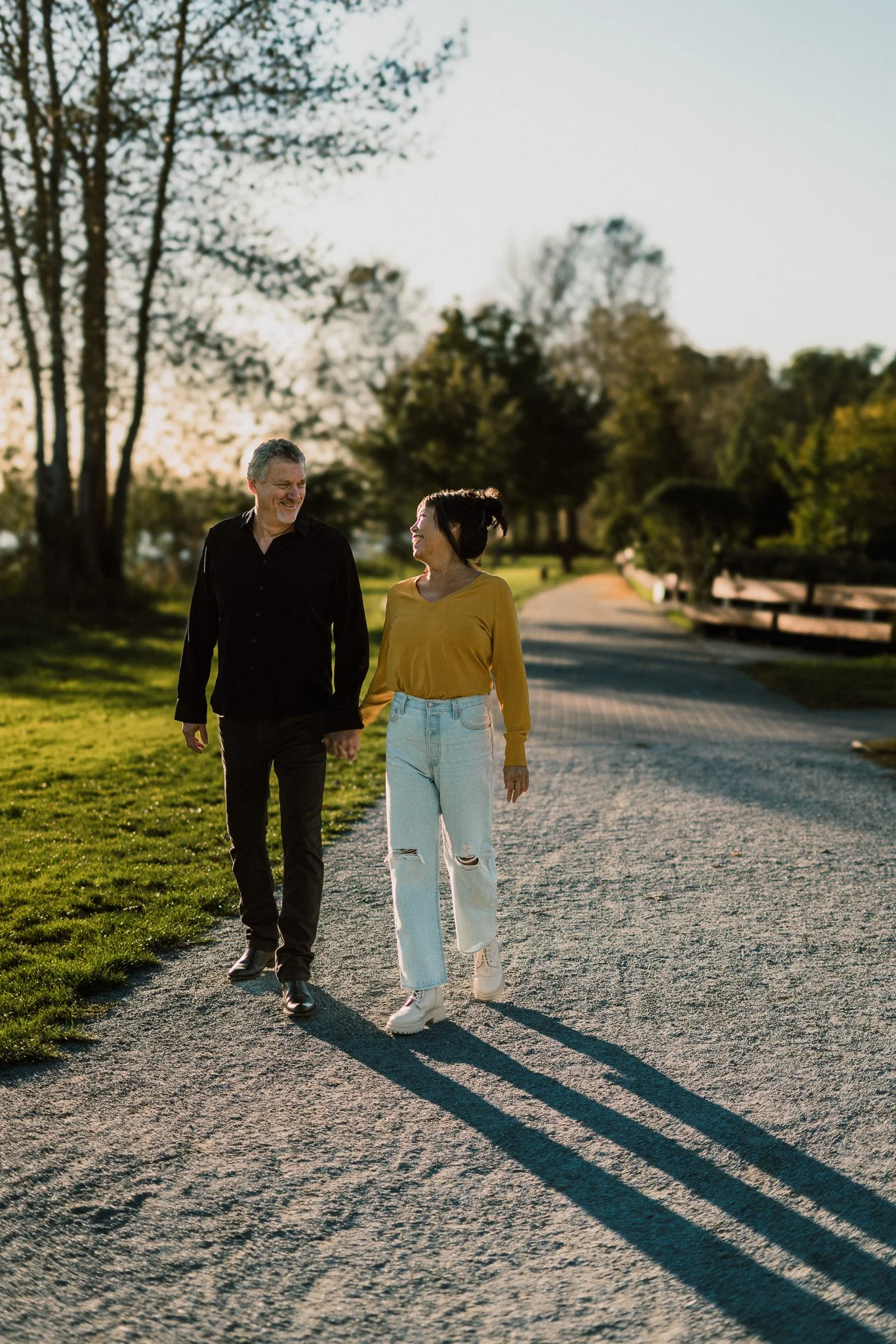 A man and a woman walking together on a gravel path in a park during sunset, smiling at each other, with trees and greenery in the background.