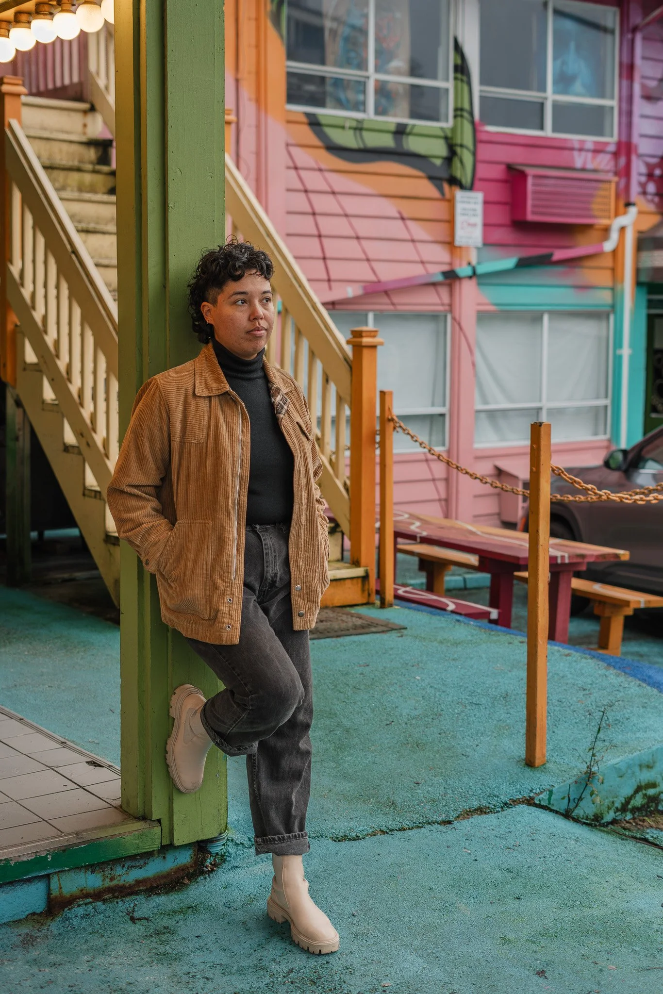 A woman with short curly hair, wearing a brown jacket, black turtleneck, gray jeans, and white boots, standing outside a colorful building with pink and orange walls, leaning against a green pillar.