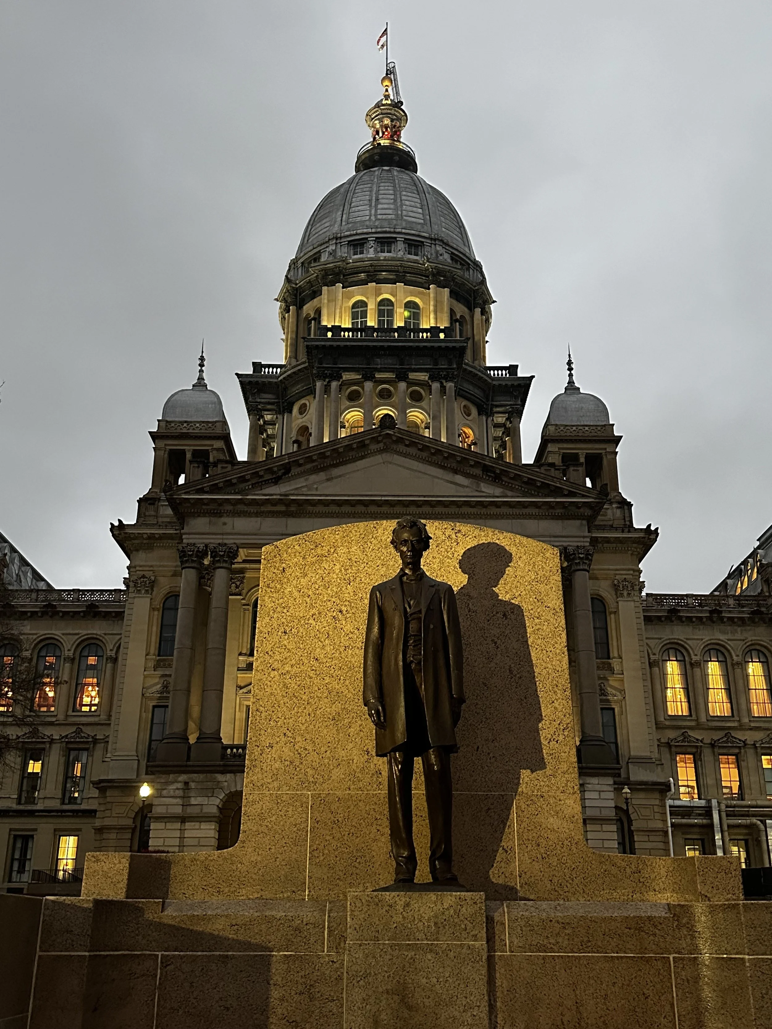 A bronze statue of Abraham Lincoln in front of the Illinois State Capitol building in Springfield, Illinois, during the evening with the building illuminated and the sky overcast.