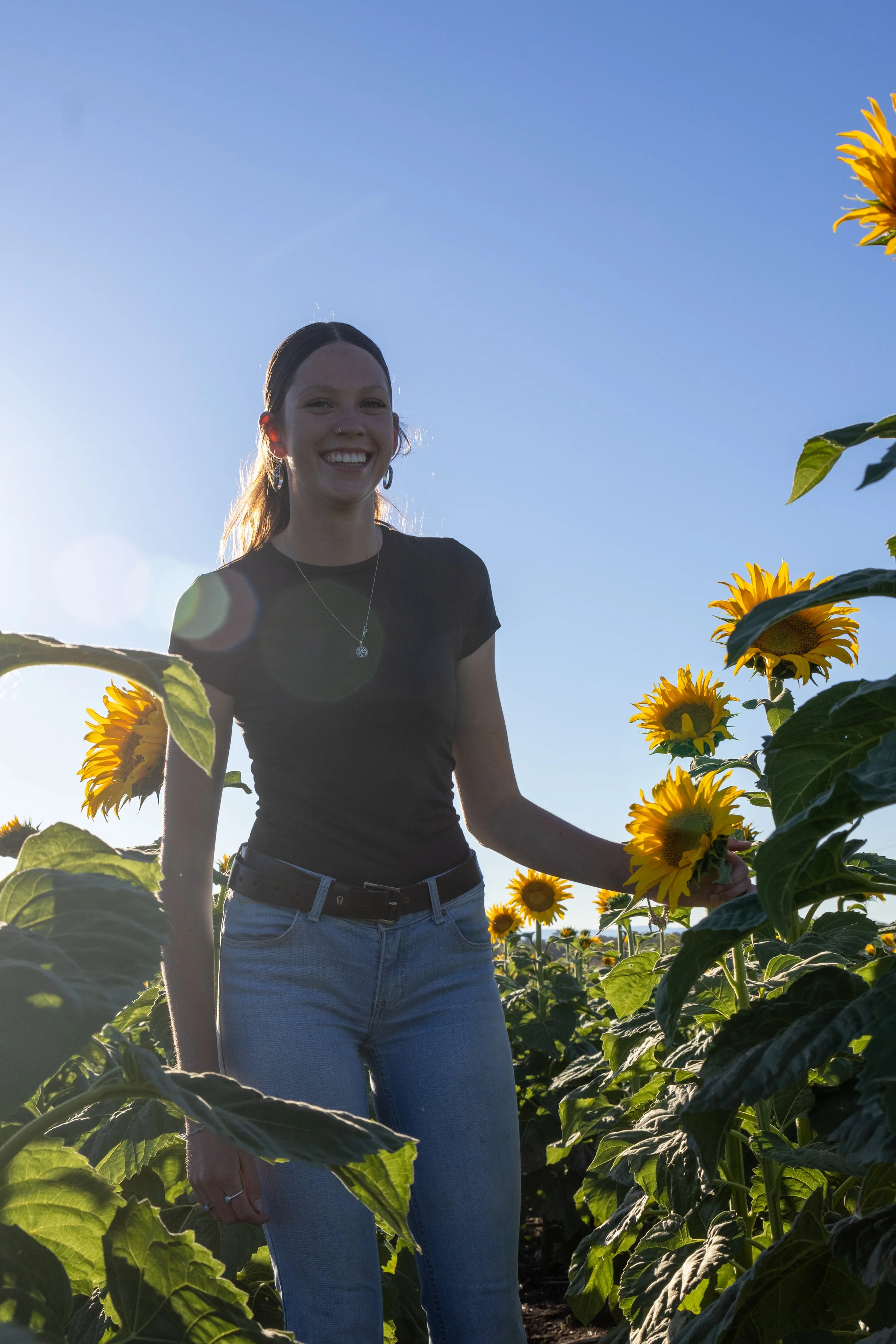 A young woman smiling in a sunflower field during daytime, sunlight behind her.