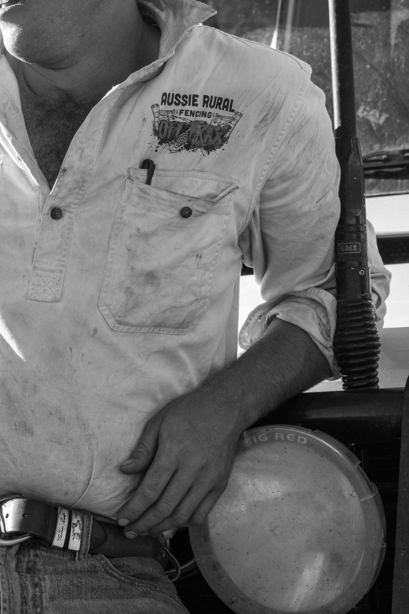 A man wearing a white shirt with an 'Aussie Rural Fencing' logo on it, sitting with his hand resting on his waist, with a pen in his shirt pocket, and a plunger next to him.
