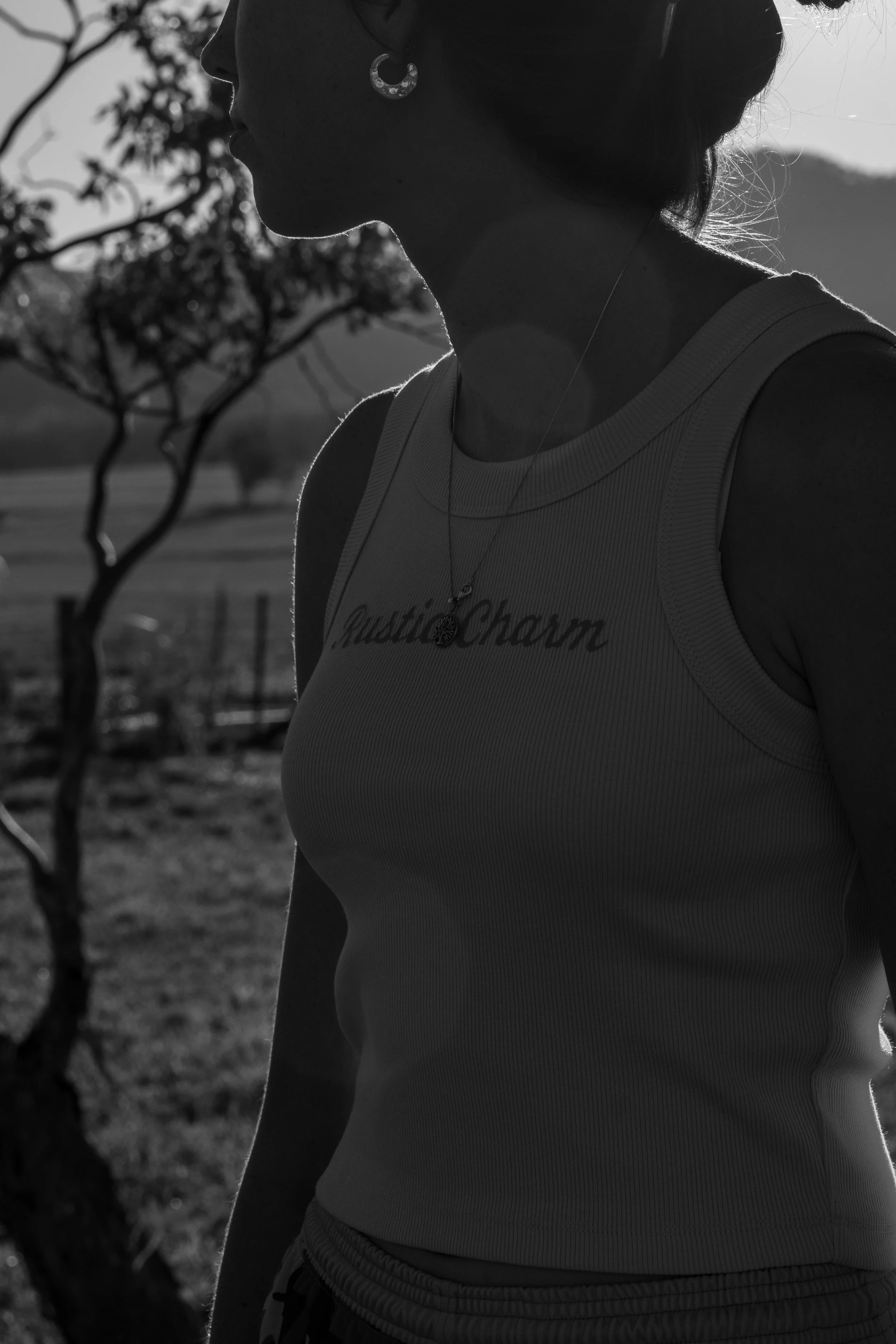 Silhouette of a woman wearing a sleeveless top and jewelry, standing outdoors with a tree and landscape in the background.