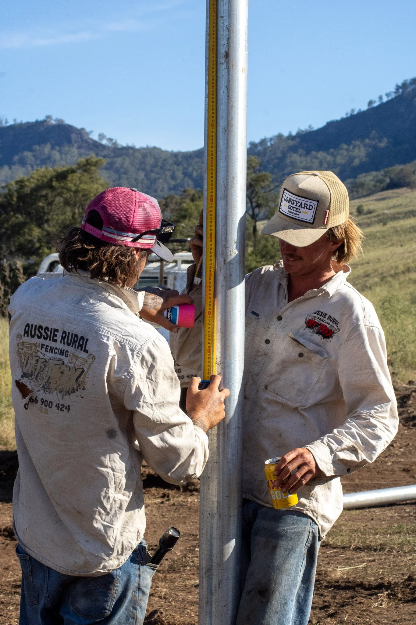 Two workers in Aussie Rural Fencing uniforms measure a metal fence post with a ruler while standing outdoors in a rural area with mountains in the background. One worker wears a pink cap and sunglasses, the other wears a beige cap, and both hold cans