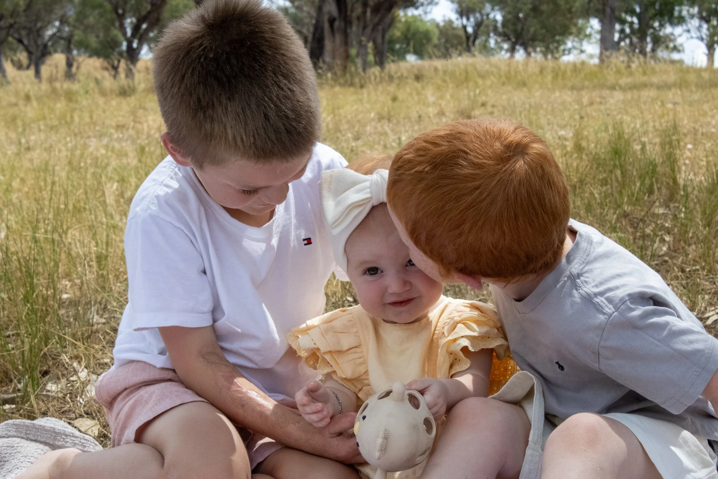 Three children outdoors, a baby girl sitting on a blanket holding a ceramic piggy bank, with two older boys leaning in towards her, in a grassy field with trees in the background.